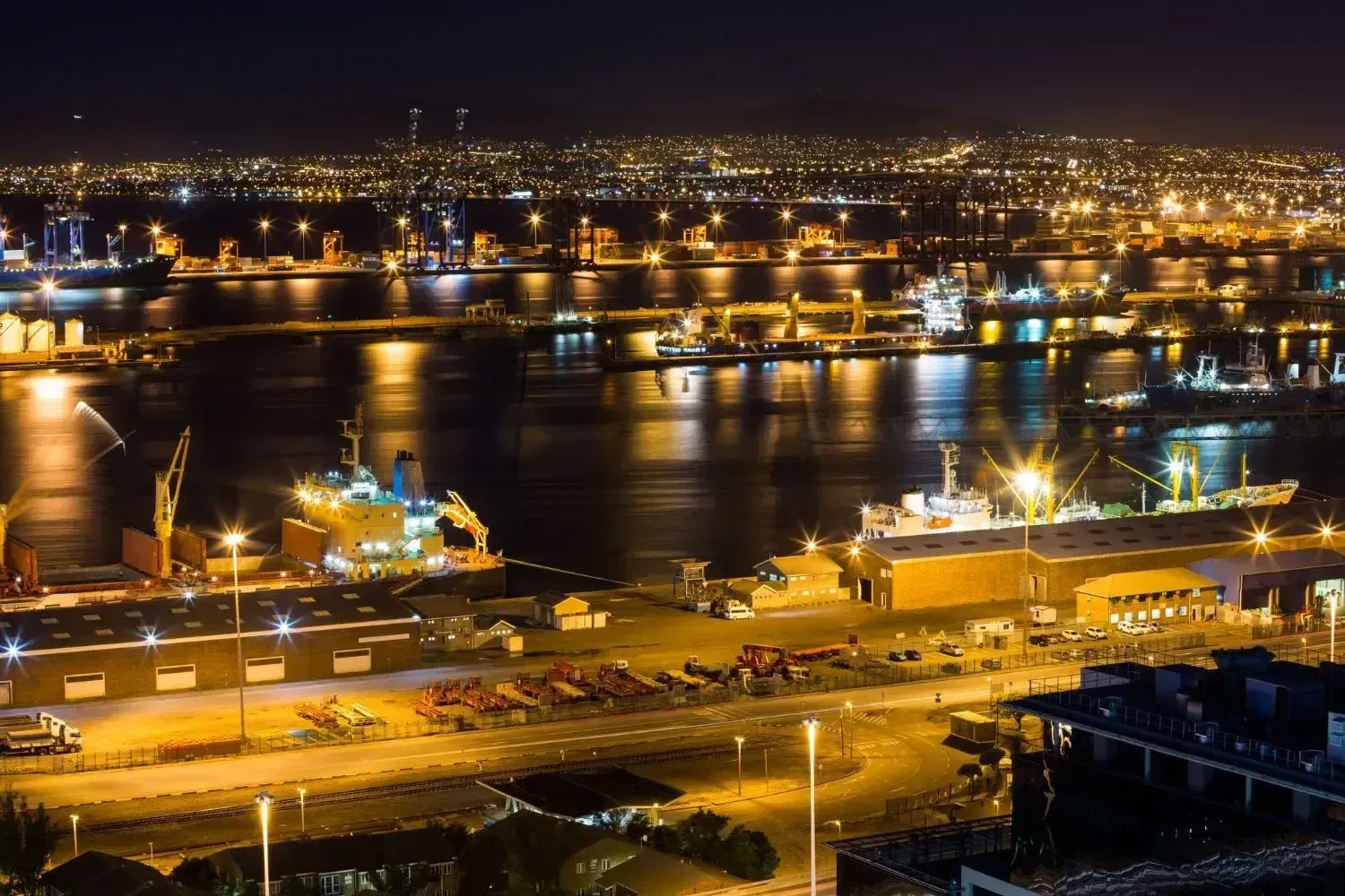 Night harbor at dusk with bright ship lights, city skyline, and reflections across the water.