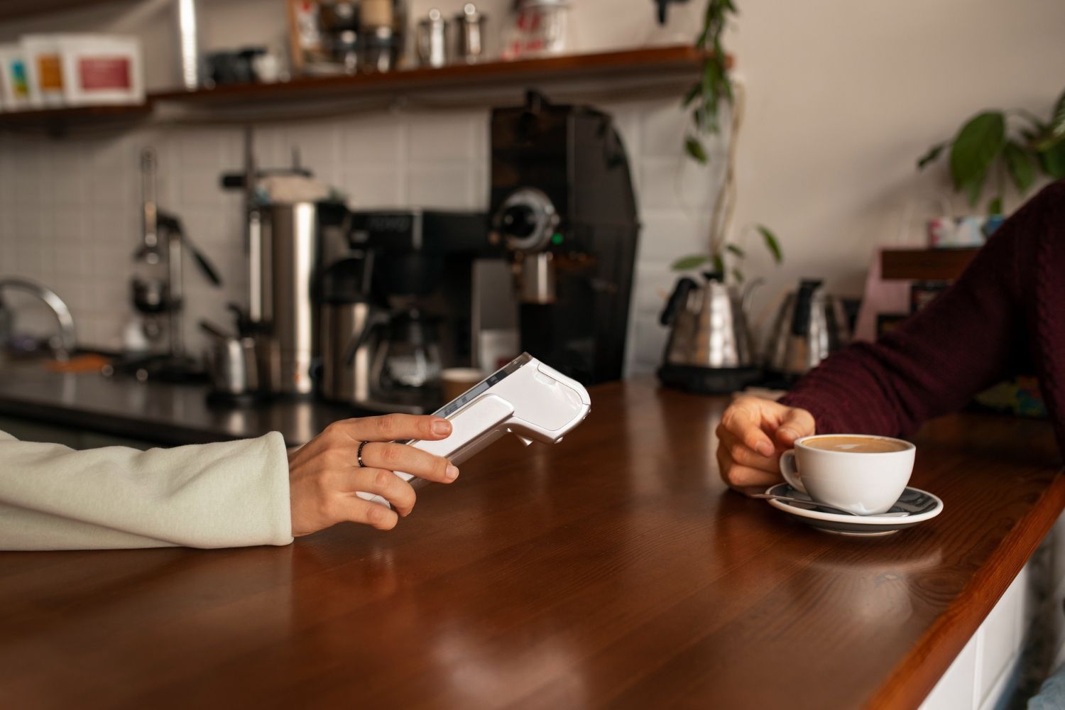 Hand pointing a white remote toward a coffee cup on a saucer at a wooden café table.