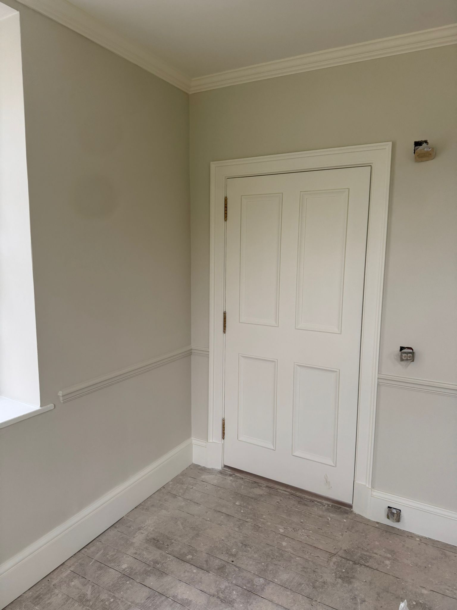 Interior corner of a room with a white door, beige walls, and crown molding. Unfinished wooden floor.