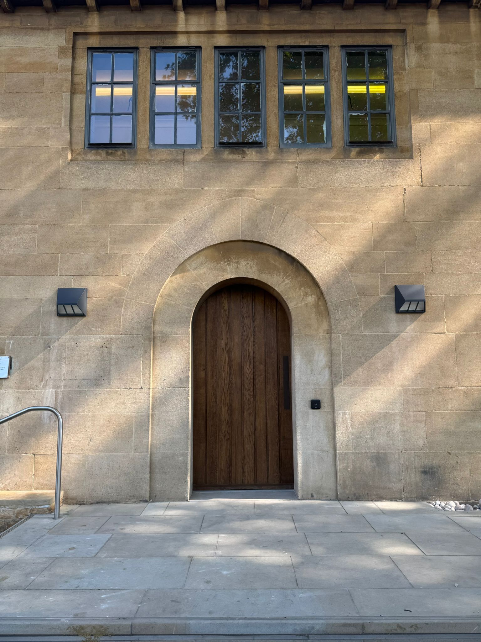 Stone building with arched wooden door, three windows above, and two square fixtures on either side of the door.
