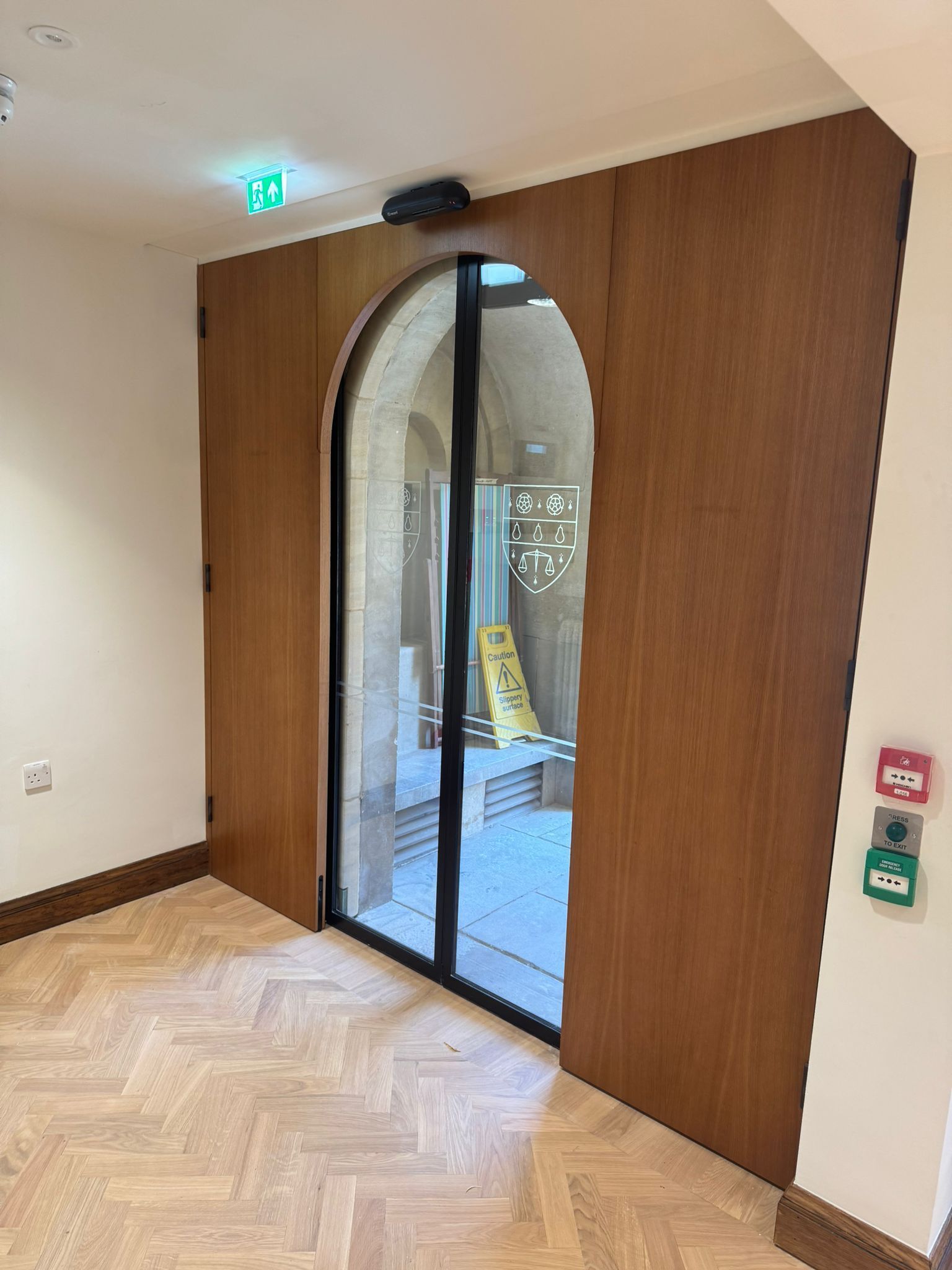 Interior view of a door with wood panels and an arched glass door. Exit sign above. Red and green buttons on wall.