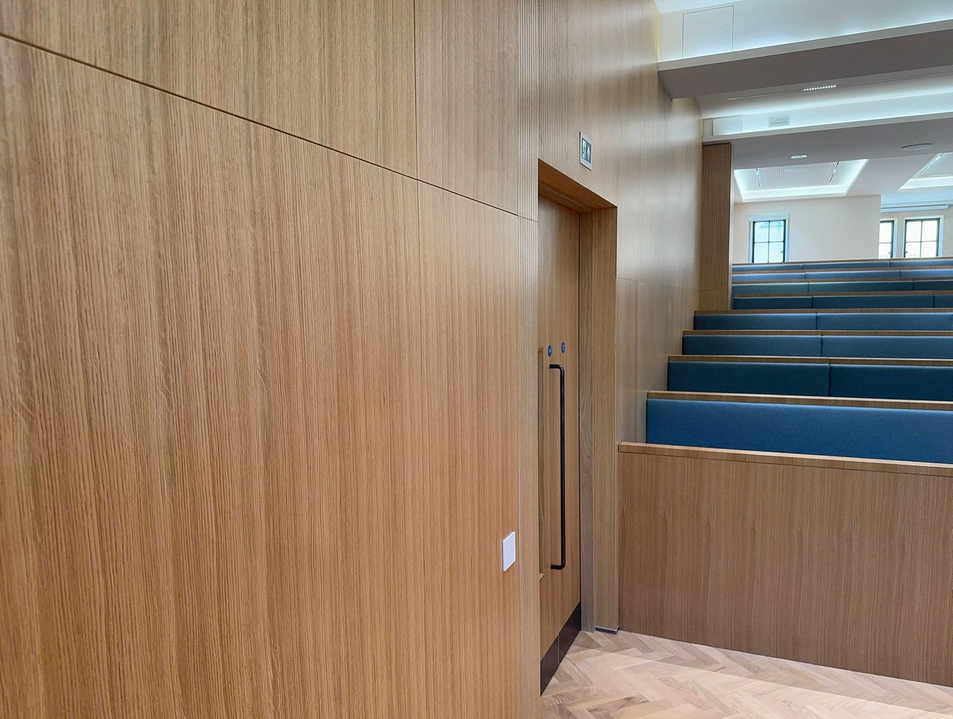 Wooden paneled lecture hall interior. Rows of blue seats. Door with handle.