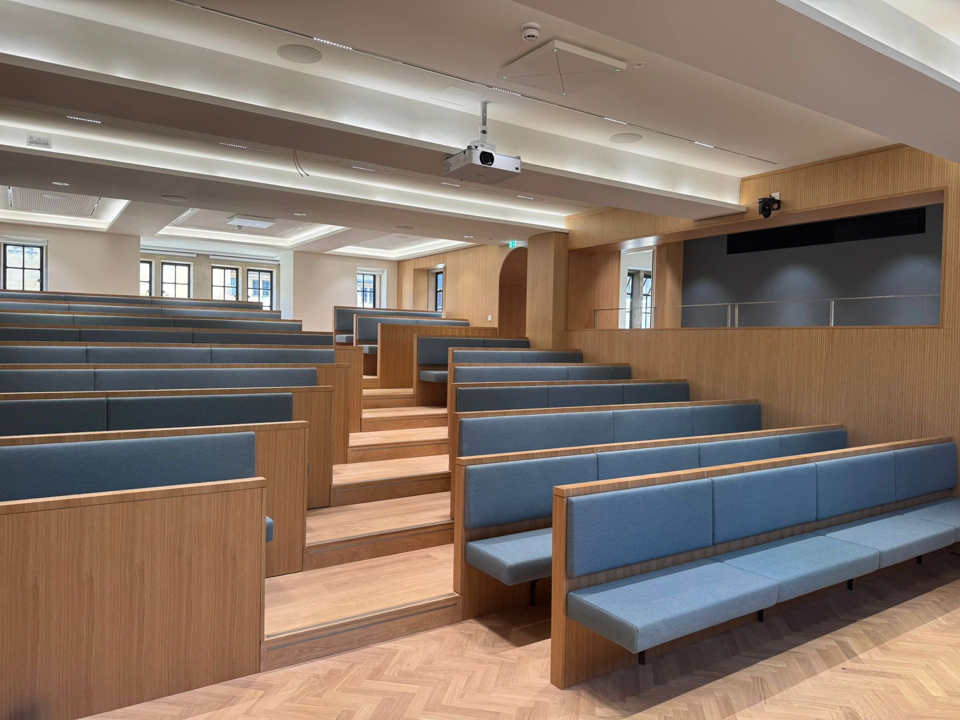 Lecture hall with tiered seating, blue benches, light wood paneling, and a projector.