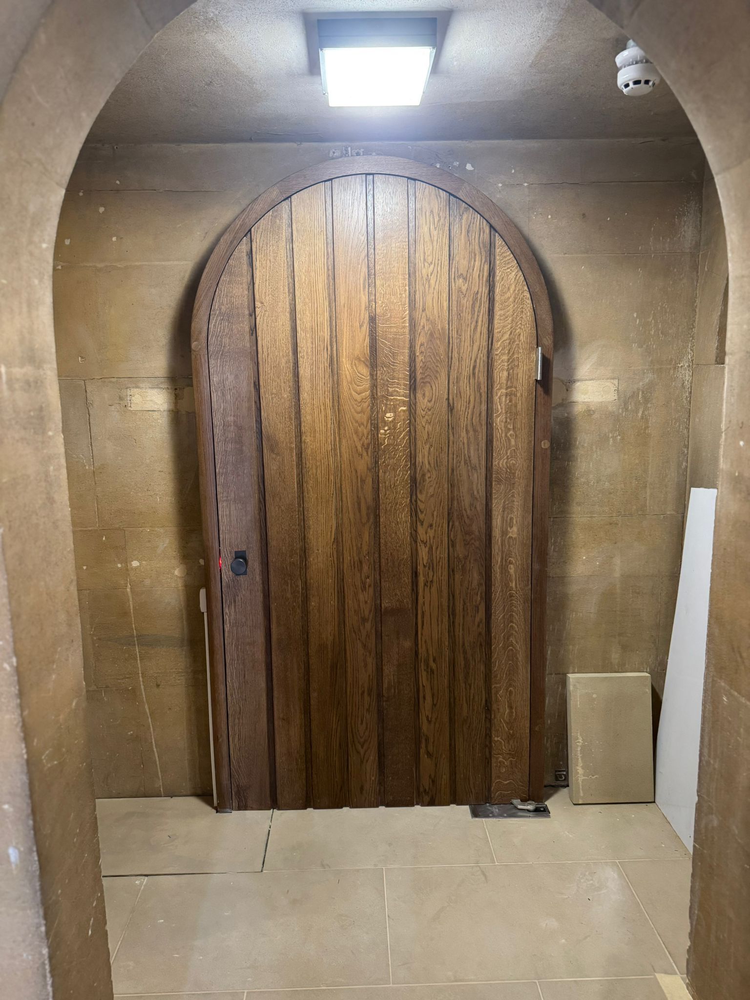 Wooden arched door in a stone-lined doorway. Lit by overhead square light.
