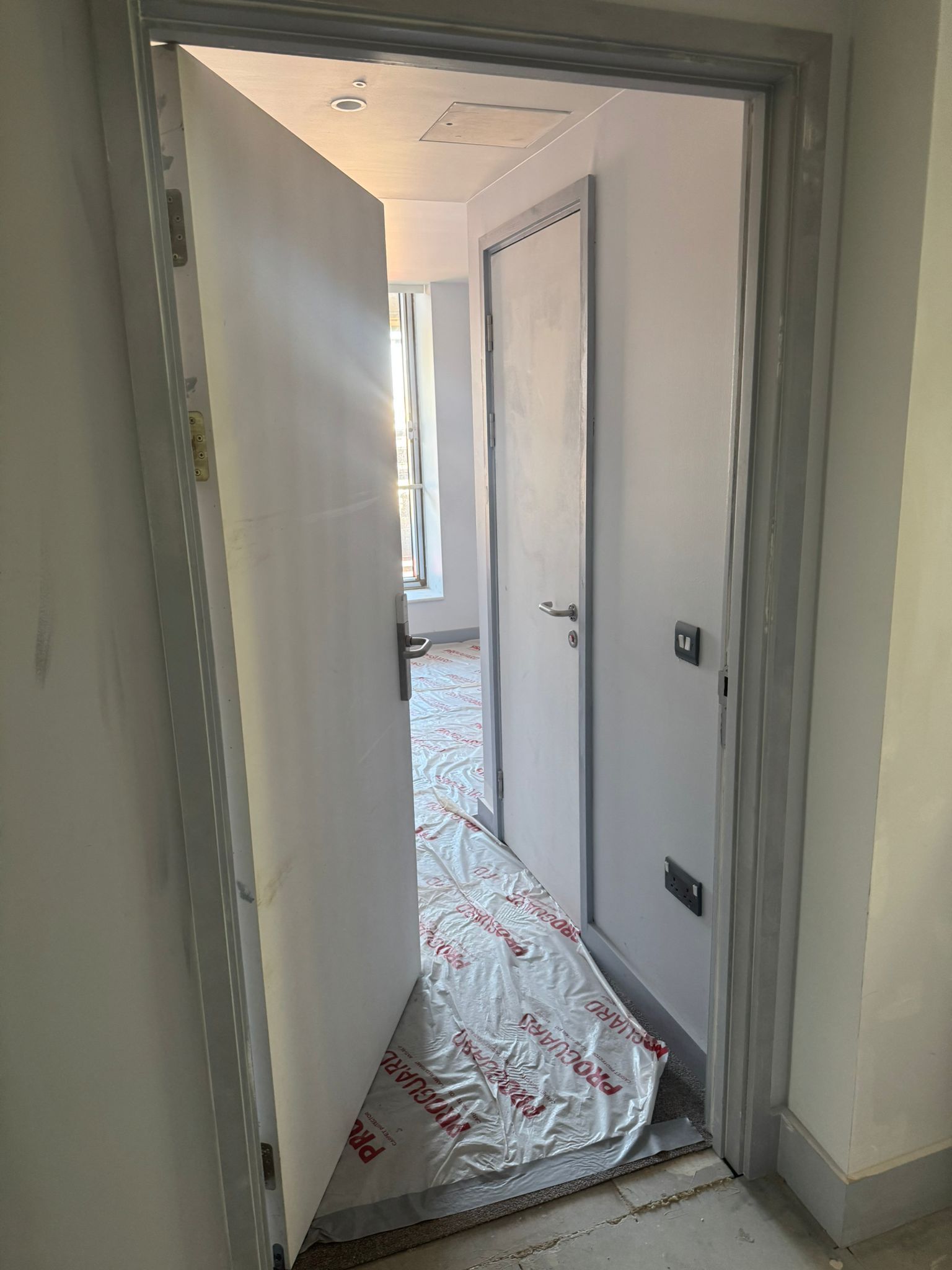 Open white door reveals a hallway with a closed door, sunlight, and a tarp on the floor.