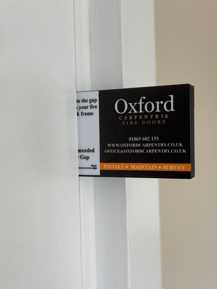 Black Oxford safety door lock label inserted between a white door and frame.