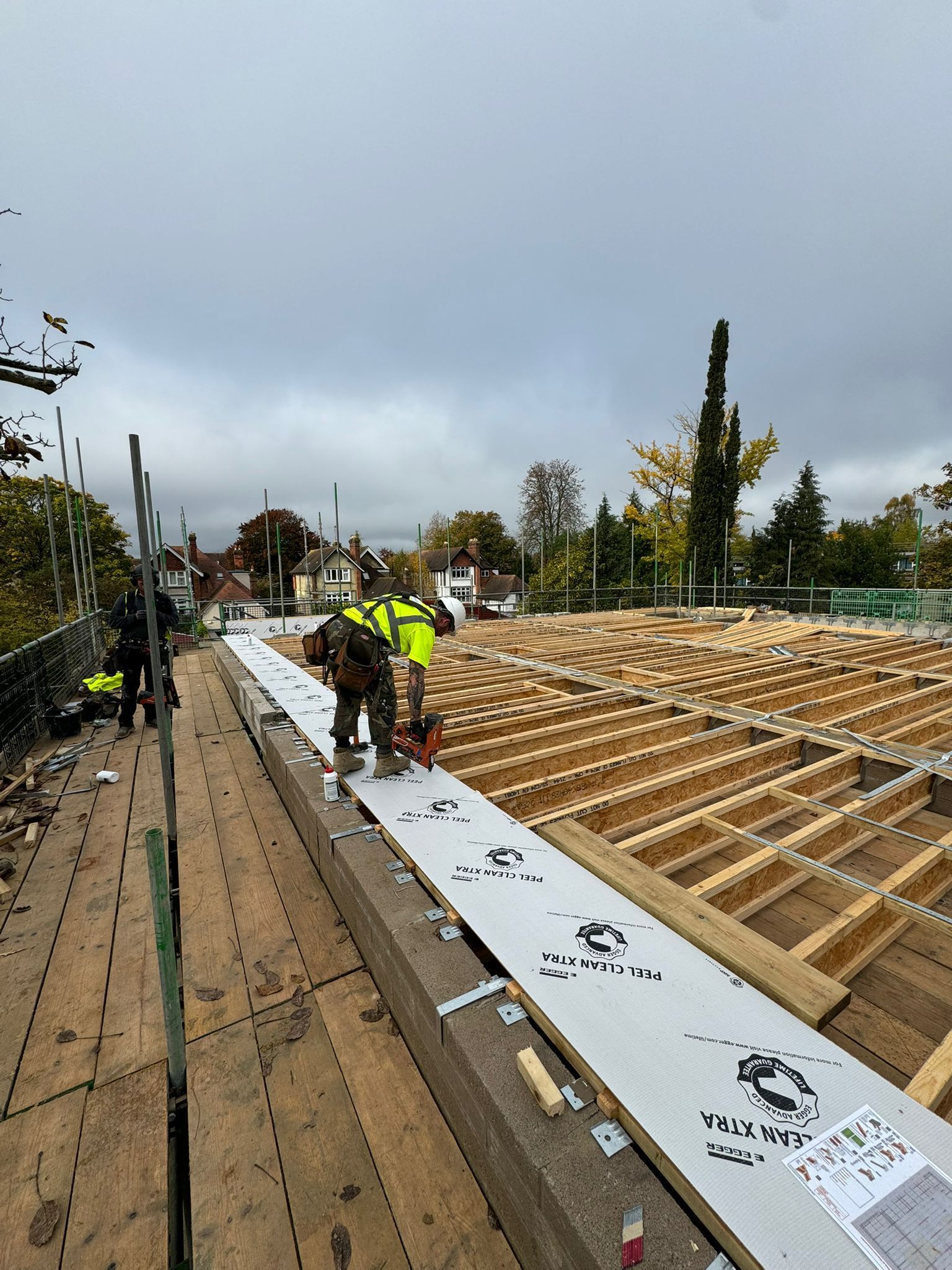 Construction workers on a partially built roof, laying down materials on wooden beams under an overcast sky.