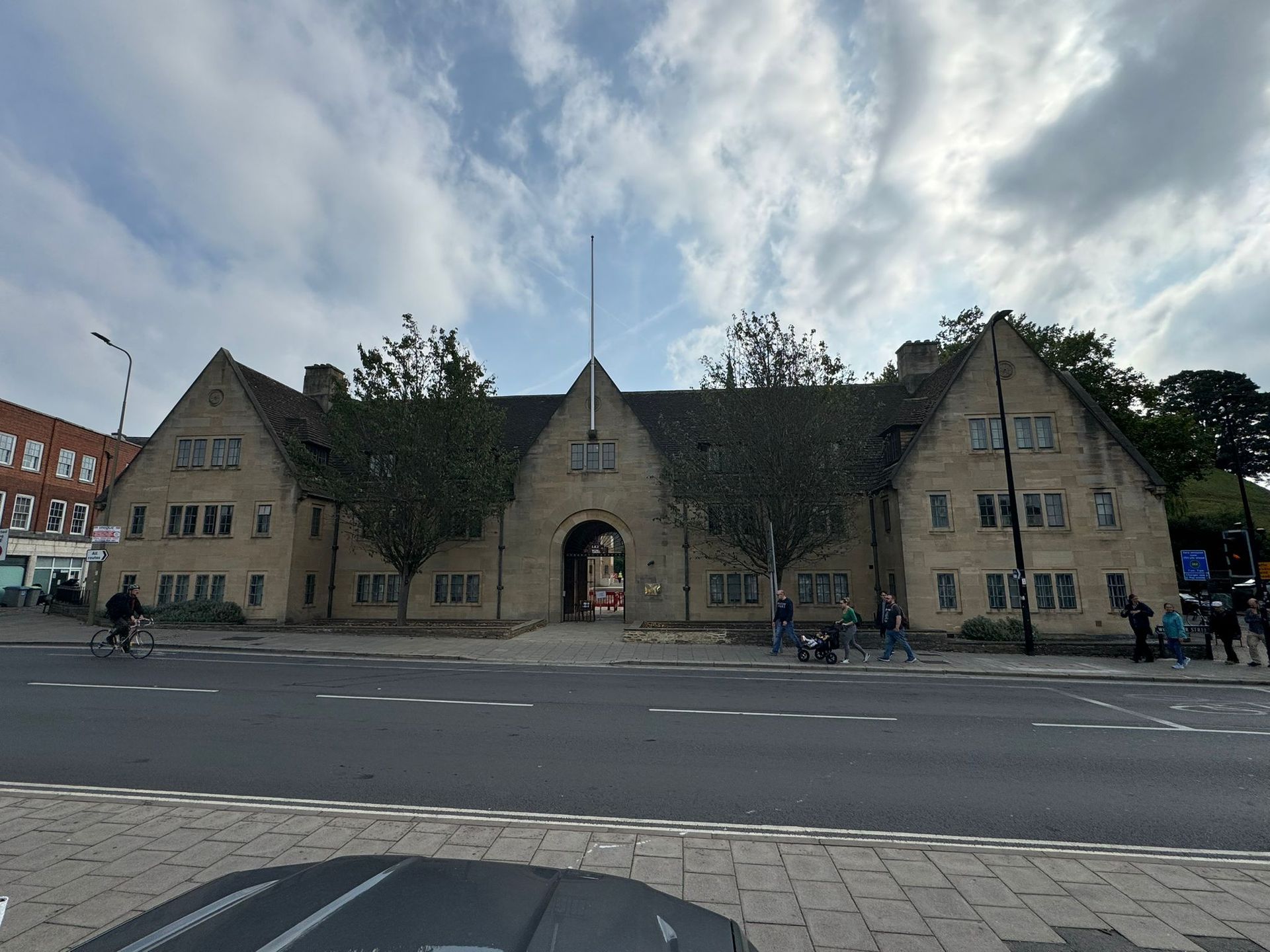 Beige building with arched entrance and gabled roof, trees, cloudy sky, people on street.