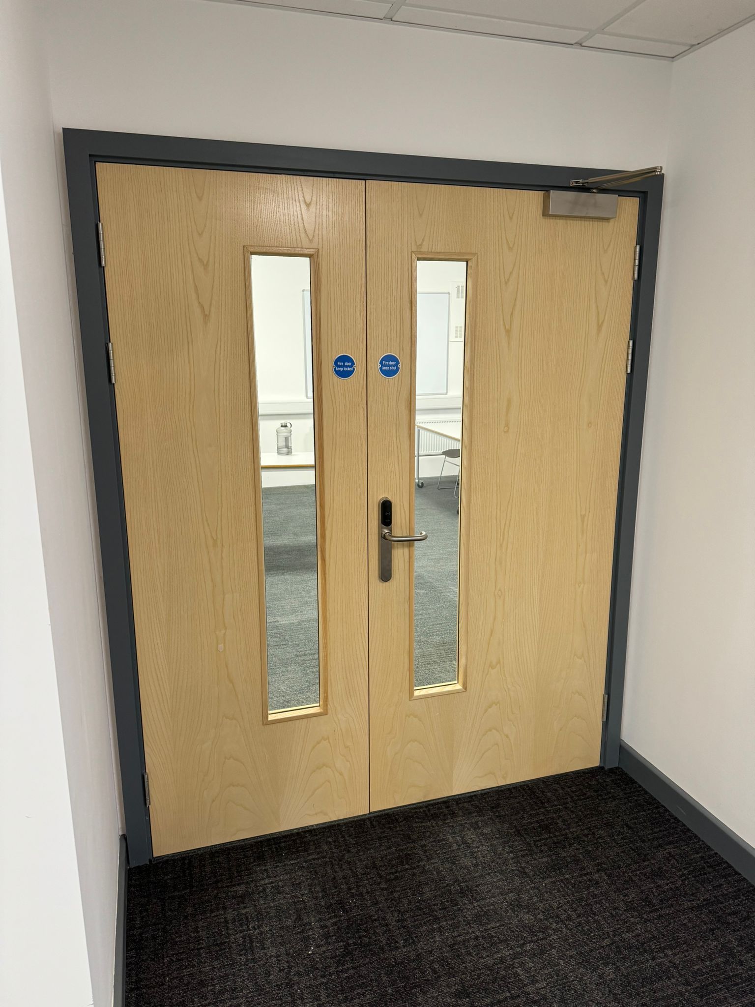 Double wooden door with two vertical windows, silver handle, and blue fire safety symbols.