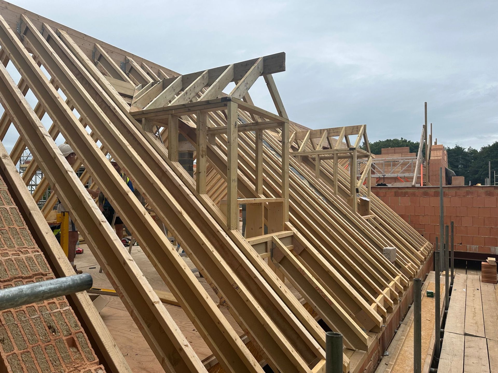 Wooden roof frame construction on a building site, with brick walls and cloudy sky.