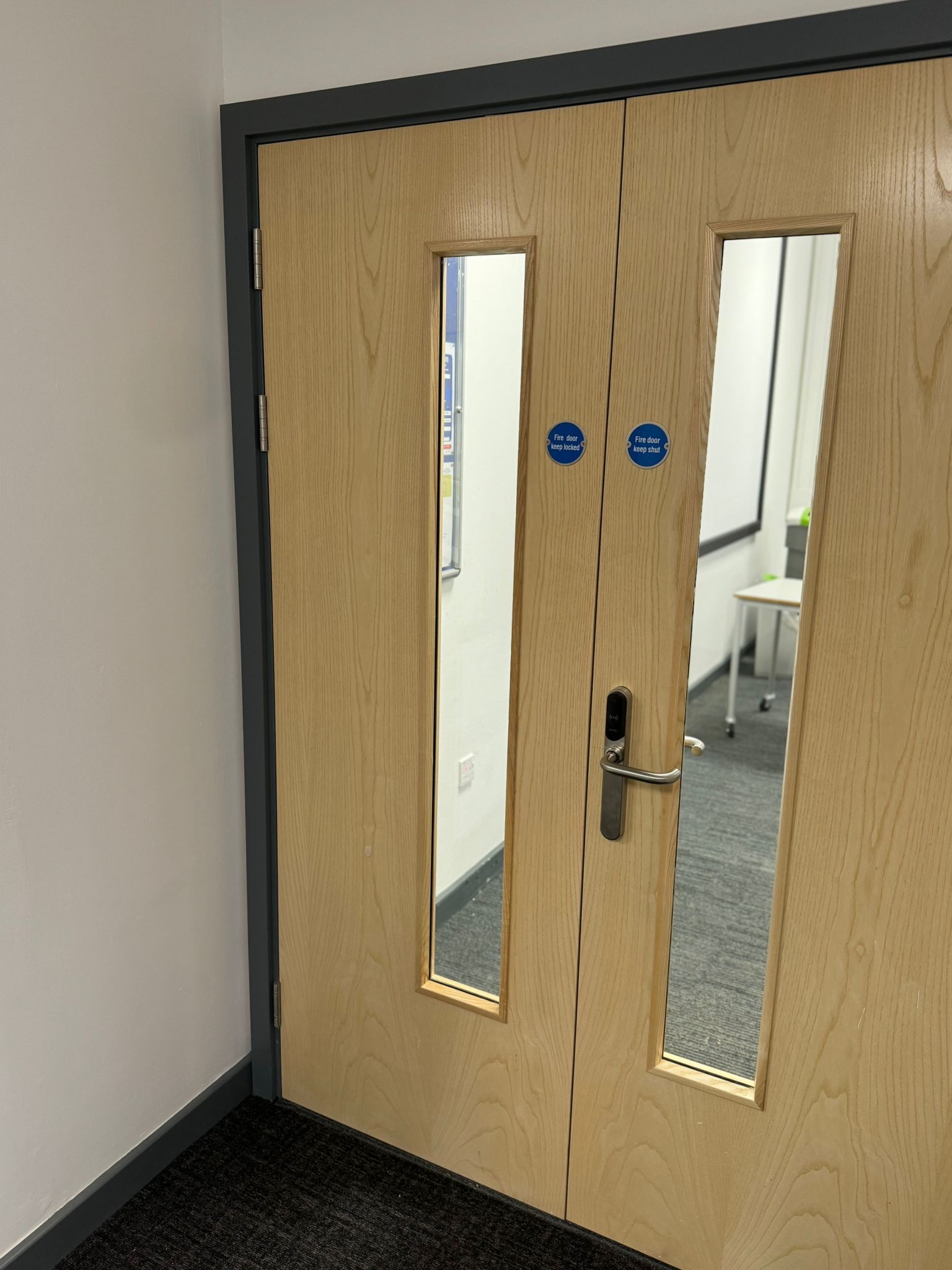 Double wooden door with rectangular windows, two fire safety symbols, and a silver handle.