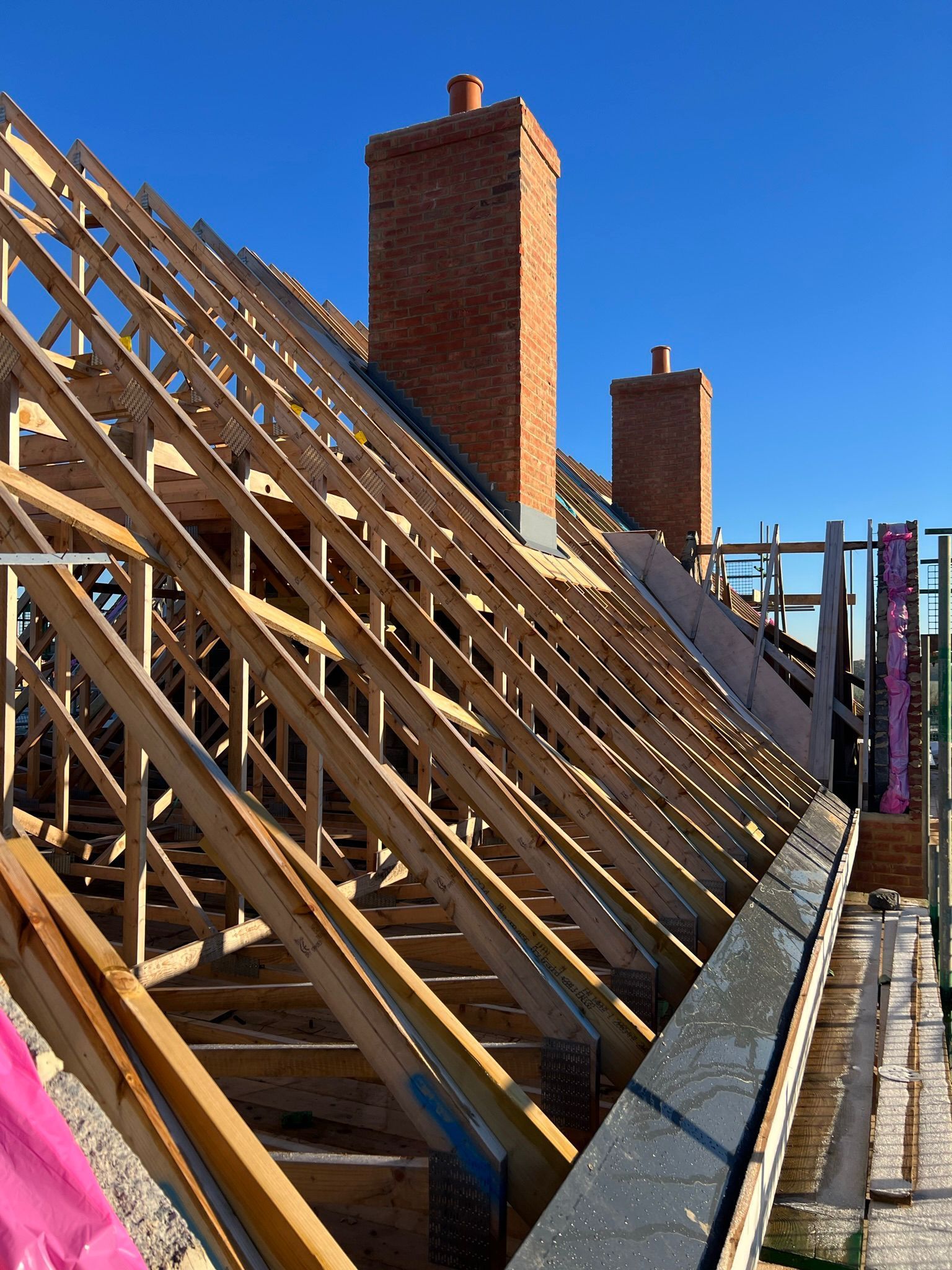Wooden roof frame under construction with brick chimneys against a clear blue sky.