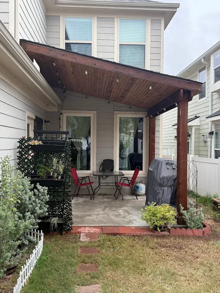 Patio with dark wooden pergola, seating, grill, and a green wall, beside a house with gray siding.