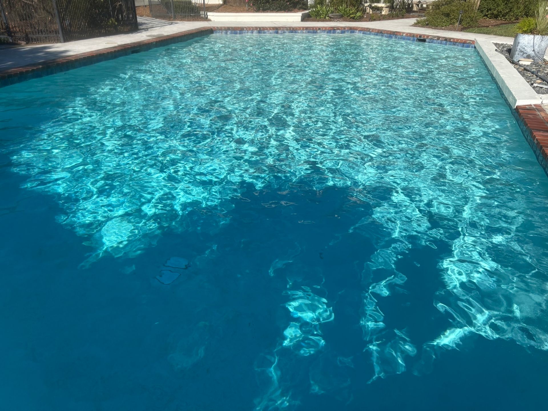 A rectangular swimming pool filled with turquoise water, reflecting sunlight with brick edging.