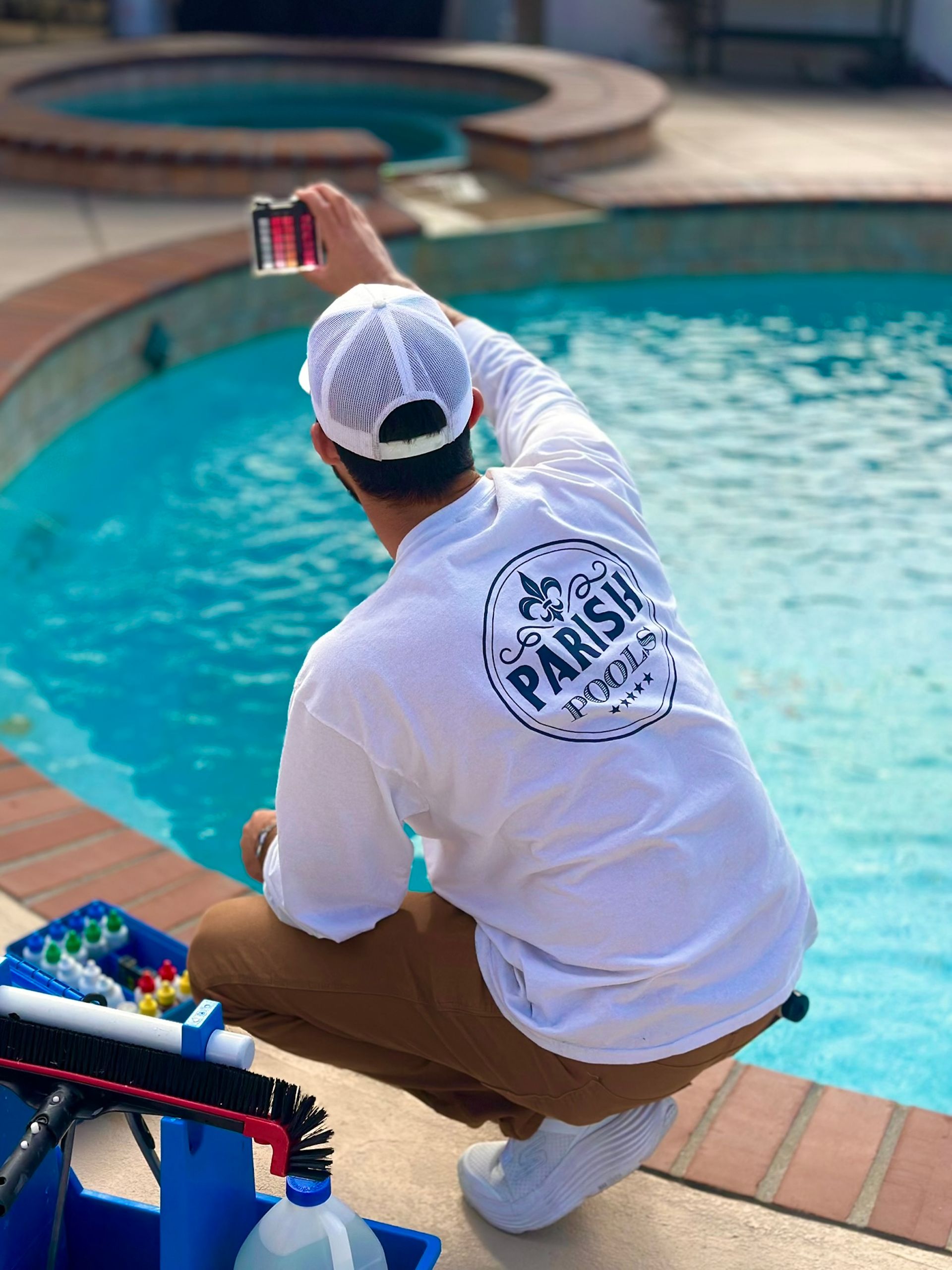 Man in white shirt with logo taking a photo of a pool with his phone. He kneels near pool's edge, holding device.