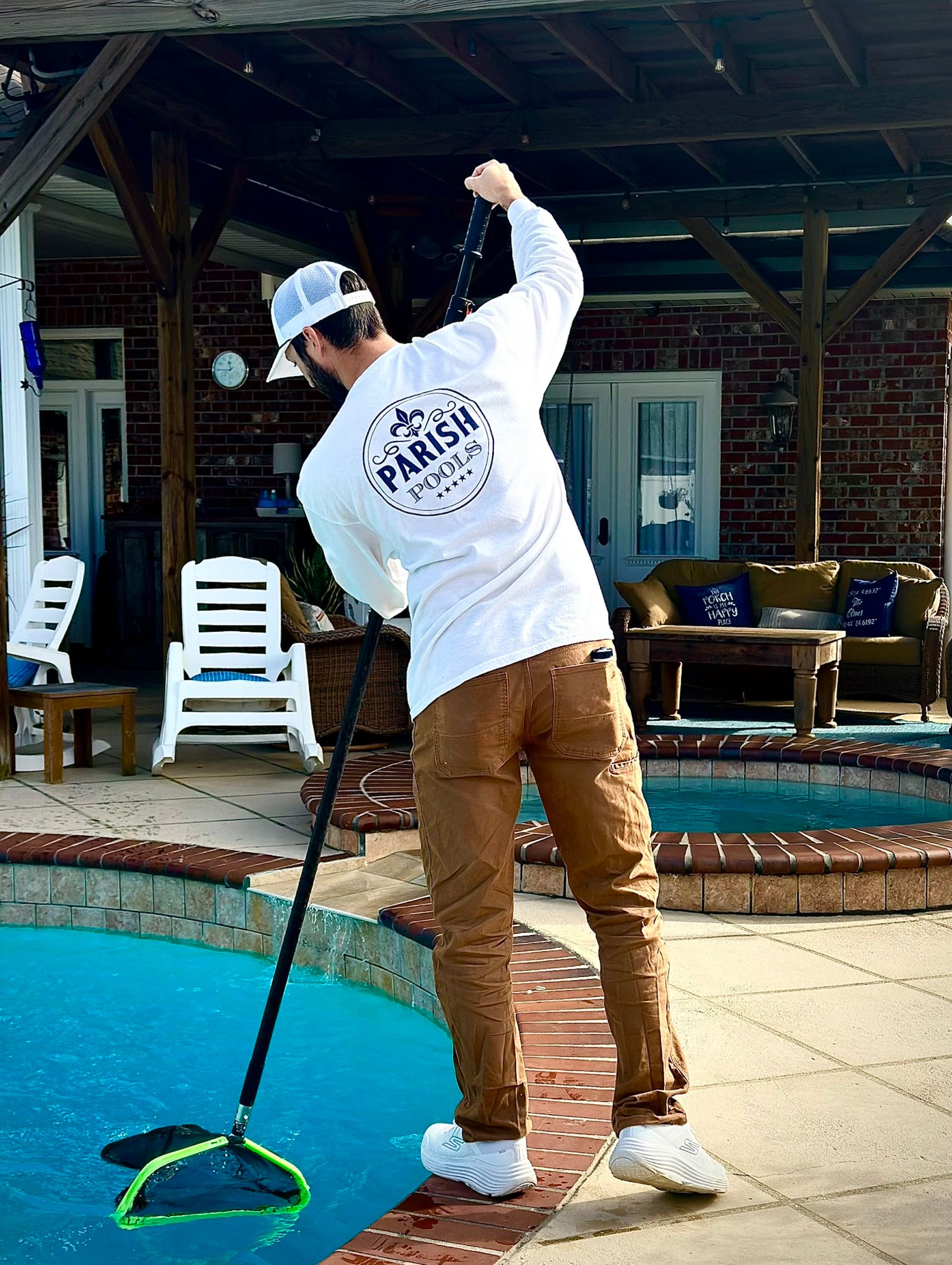Man in white shirt cleans a pool with a net. He wears a hat and brown pants. Poolside setting.