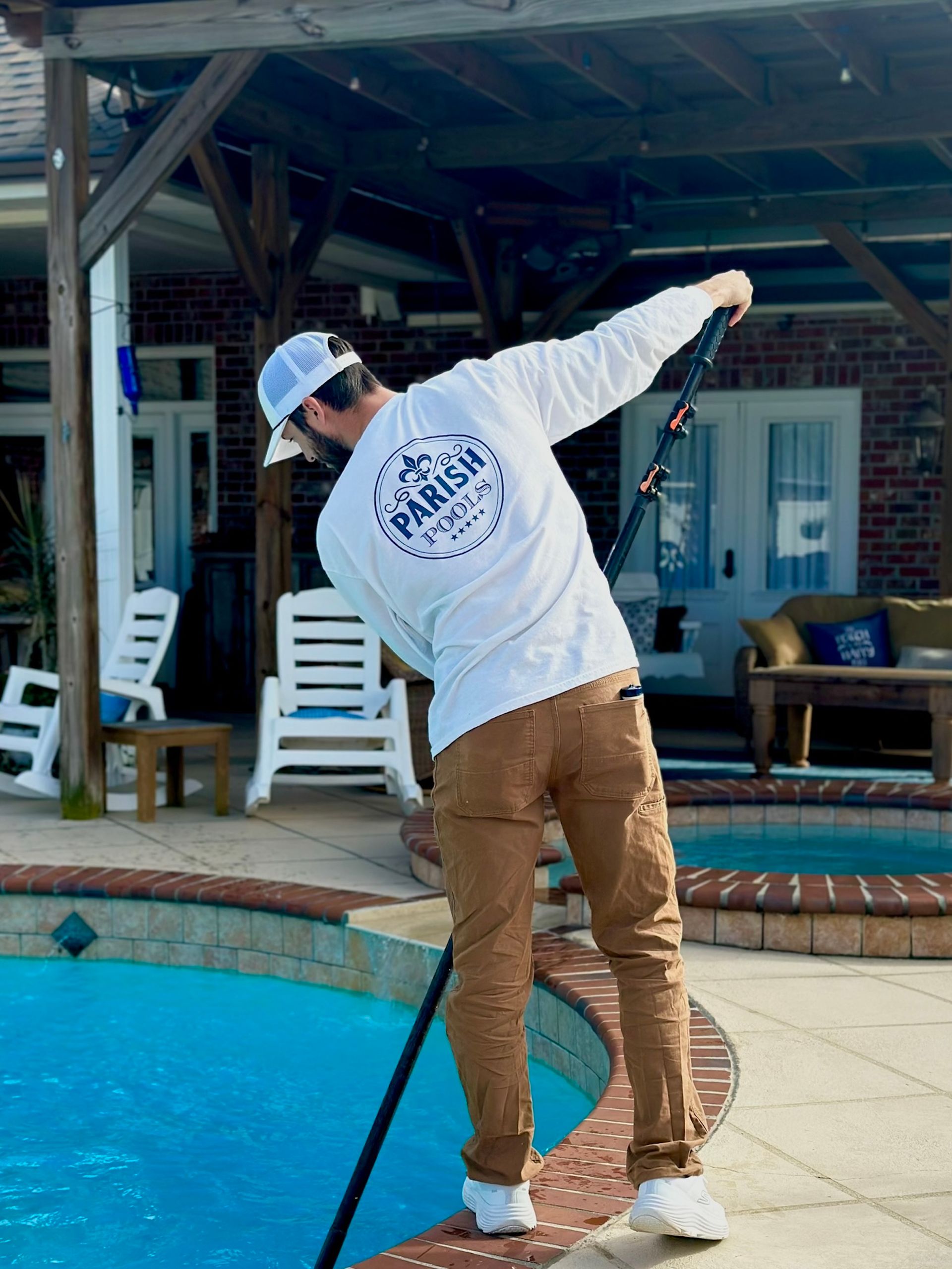 Man in white shirt and hat cleaning a pool with brown pants on a sunny day.