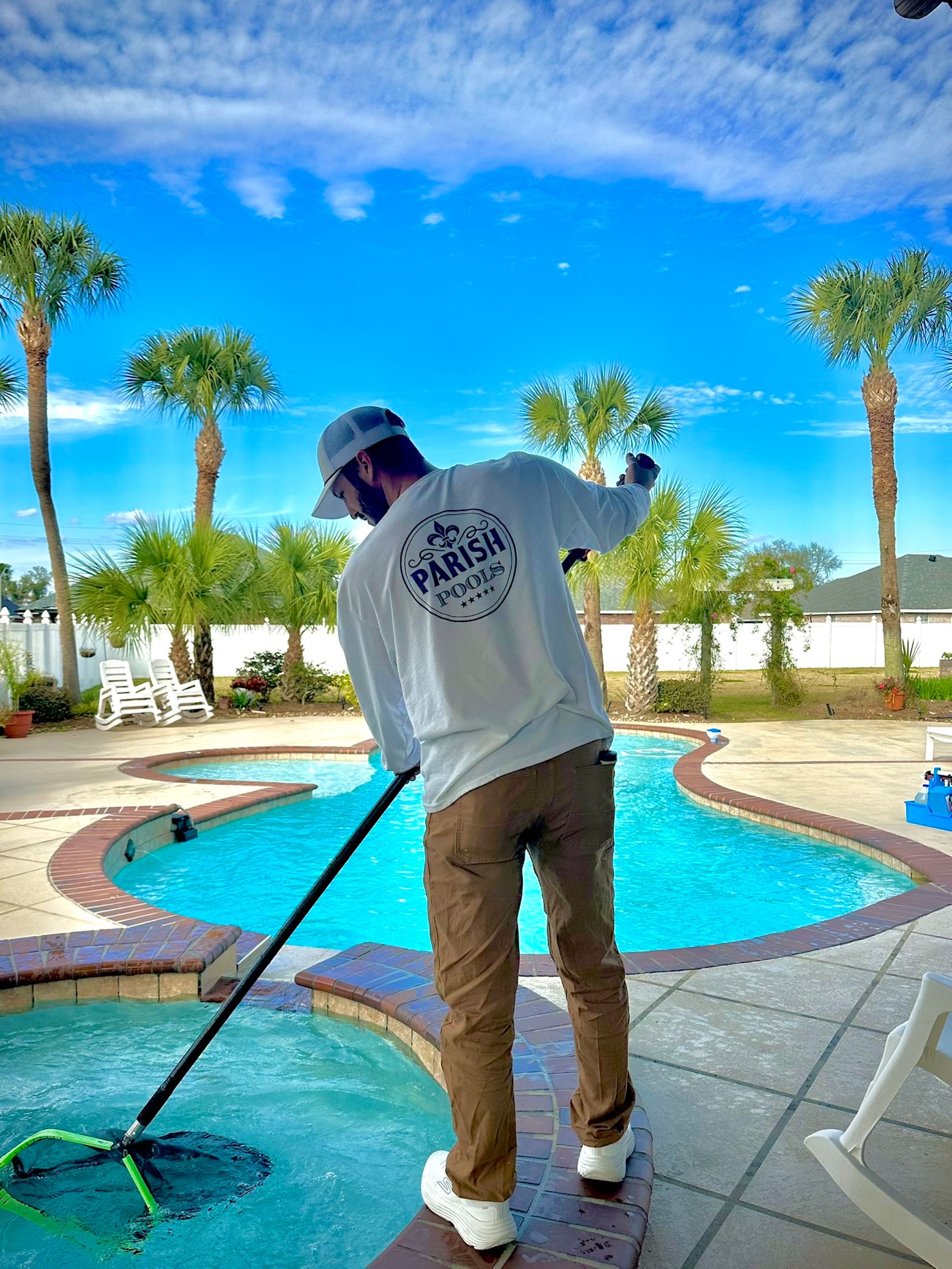 Person in long-sleeved shirt cleaning a pool with a net under a blue sky with palm trees.