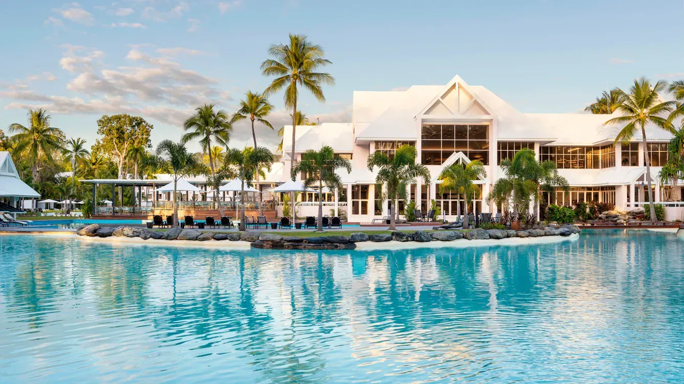 A White Resort Building Next to a Large Blue Pool With Palm Trees — Pete's Painting Solutions In Cairns, QLD