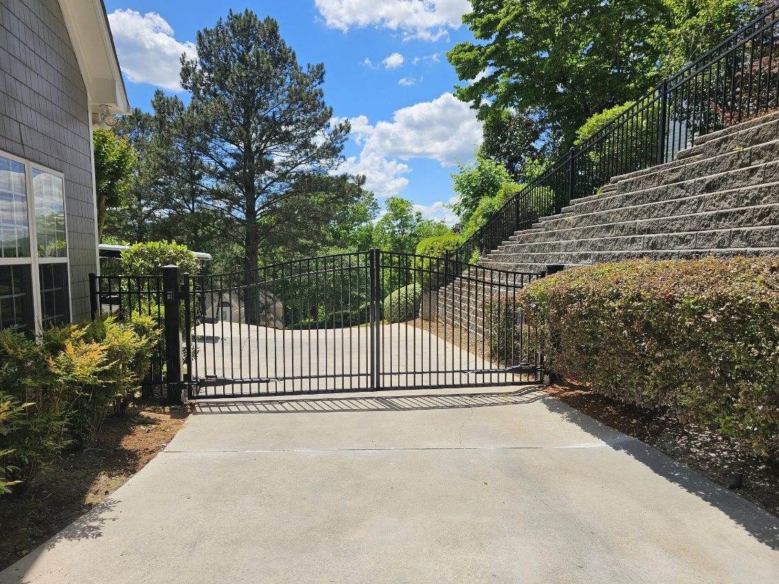 Closed black metal gate at the end of a driveway between a building and stone steps under a sunny sky