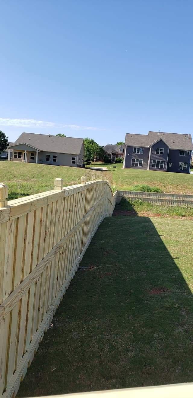 Wooden fence beside a grassy yard, with suburban houses under a clear blue sky.