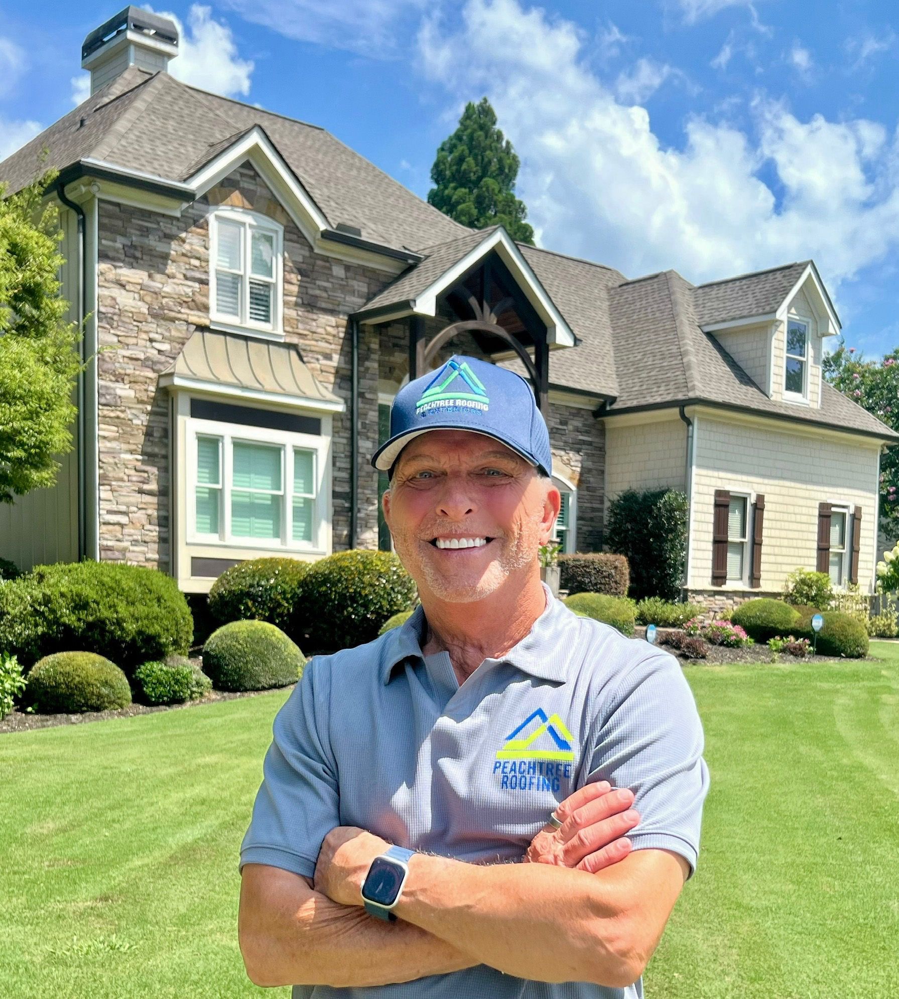 Man in front of a house, arms crossed. He wears a blue hat and a gray shirt with a logo. Green lawn and blue sky.