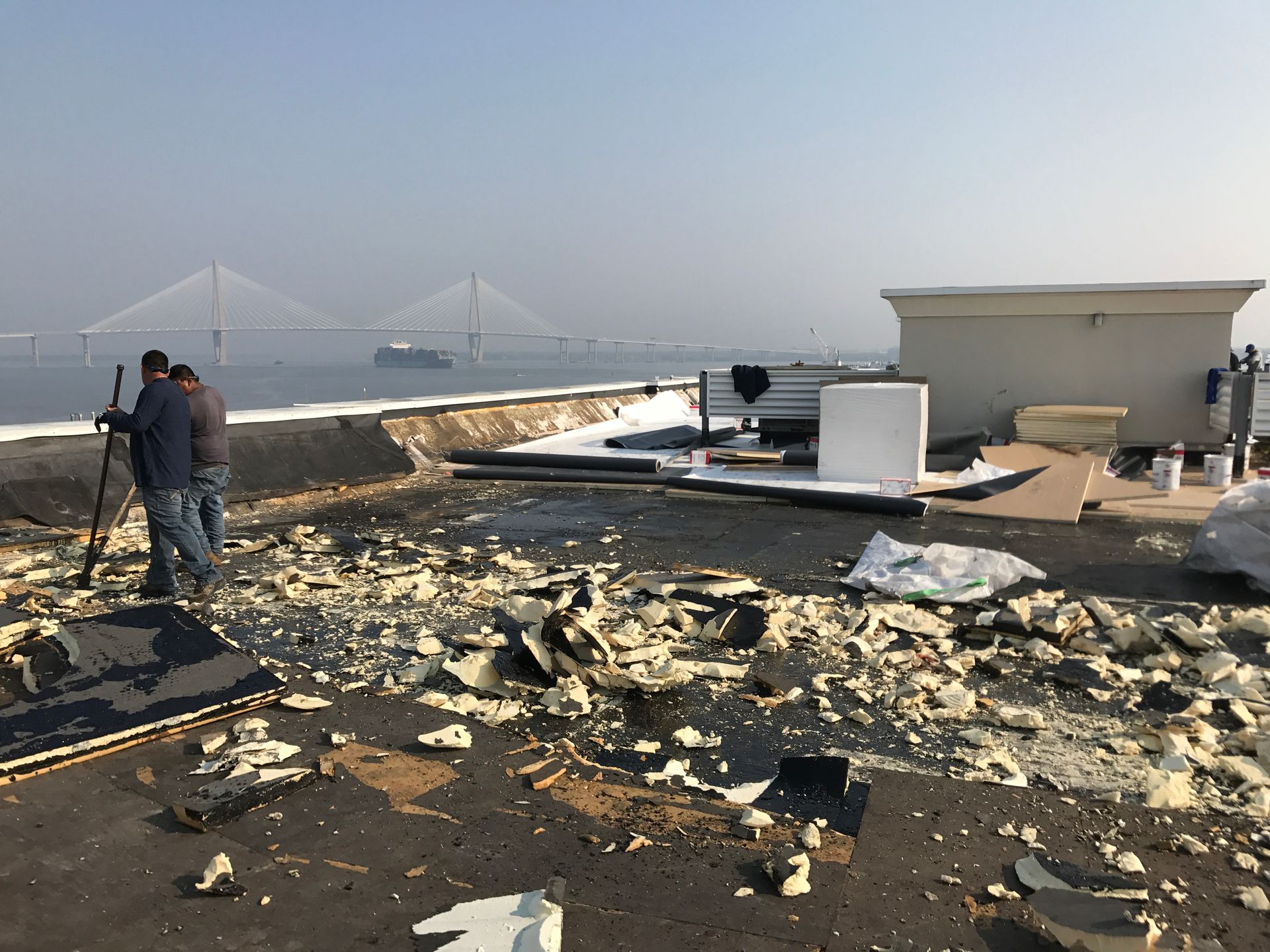 Two people on a rooftop with construction debris, bridge and water in background.