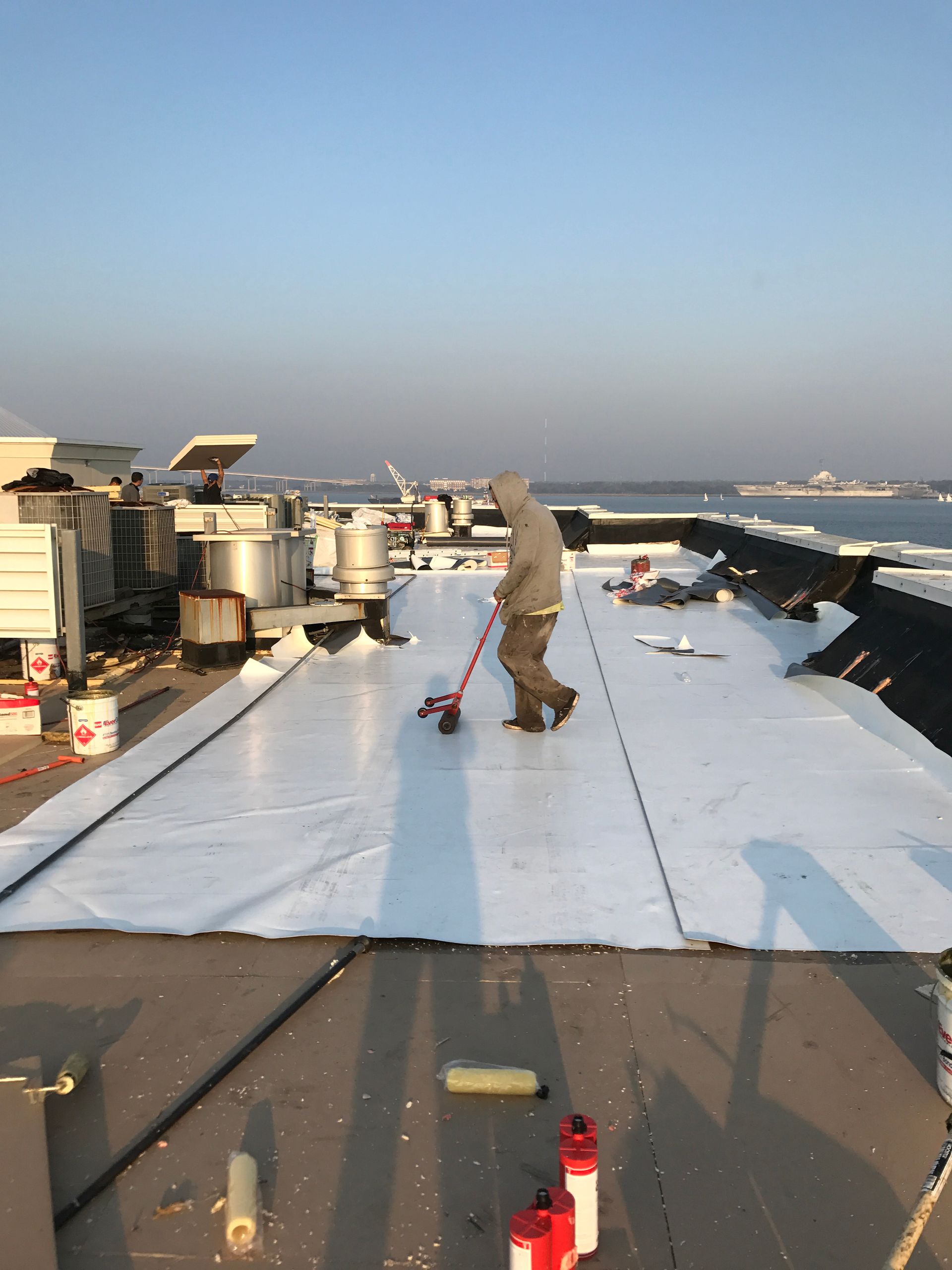 Person on a flat rooftop using a tool on a white sheet. Other equipment, buildings, and water in the distance.