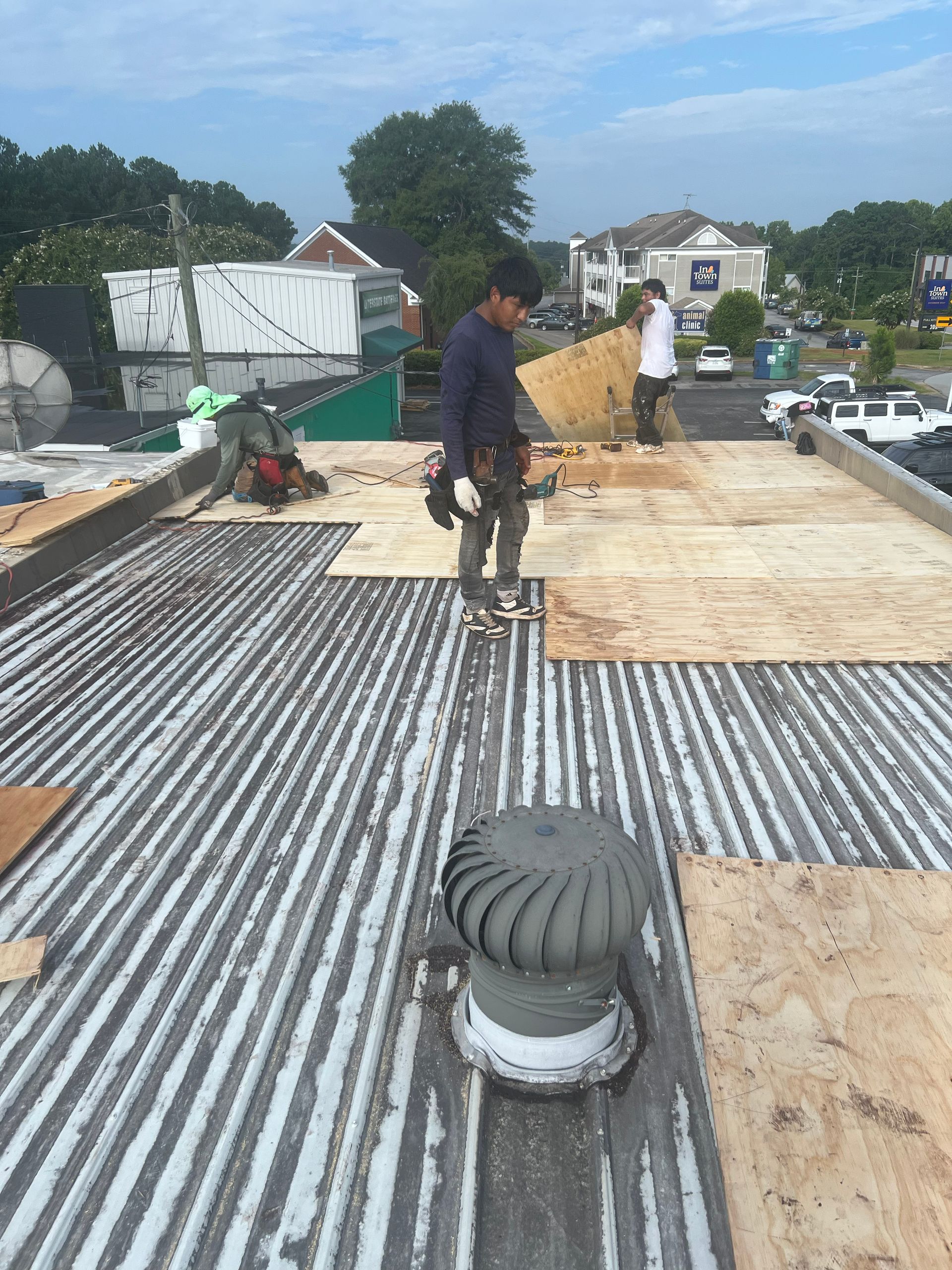 Workers on a commercial roof installing plywood over corrugated metal. Cloudy sky.