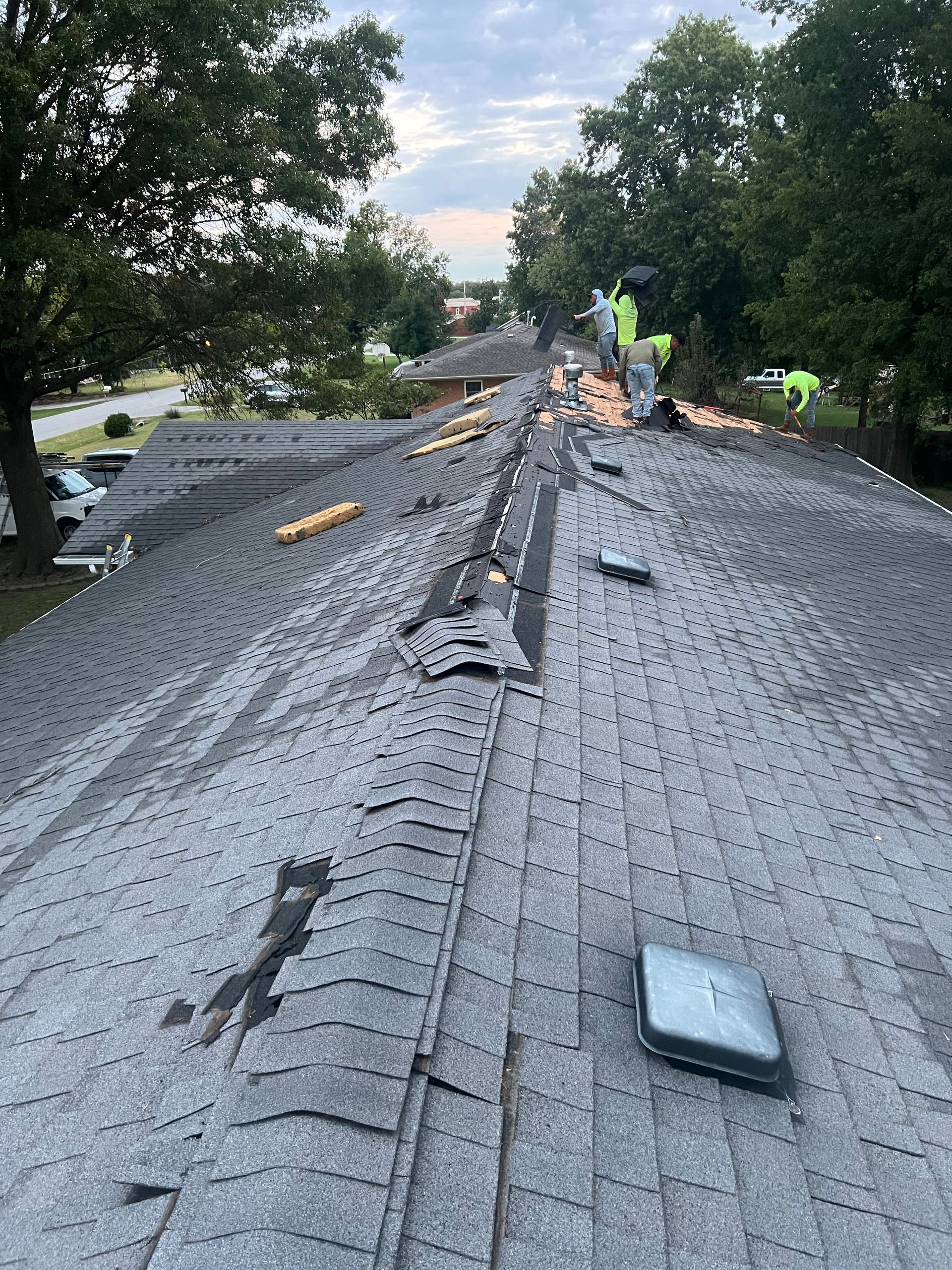 Roofing crew repairing a gray asphalt shingle roof. The roof is partially torn up. Trees and sky in the background.