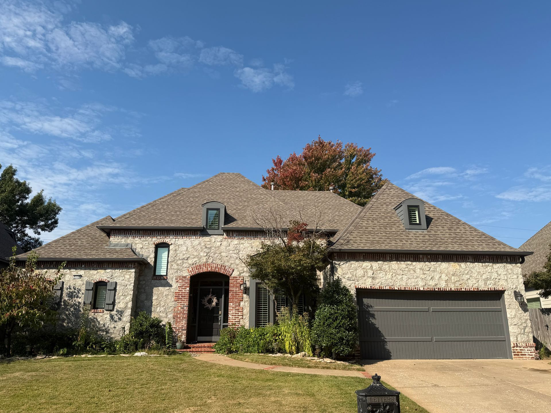 Stone-clad house with gray garage door, red door, and shingled roof under a blue sky.