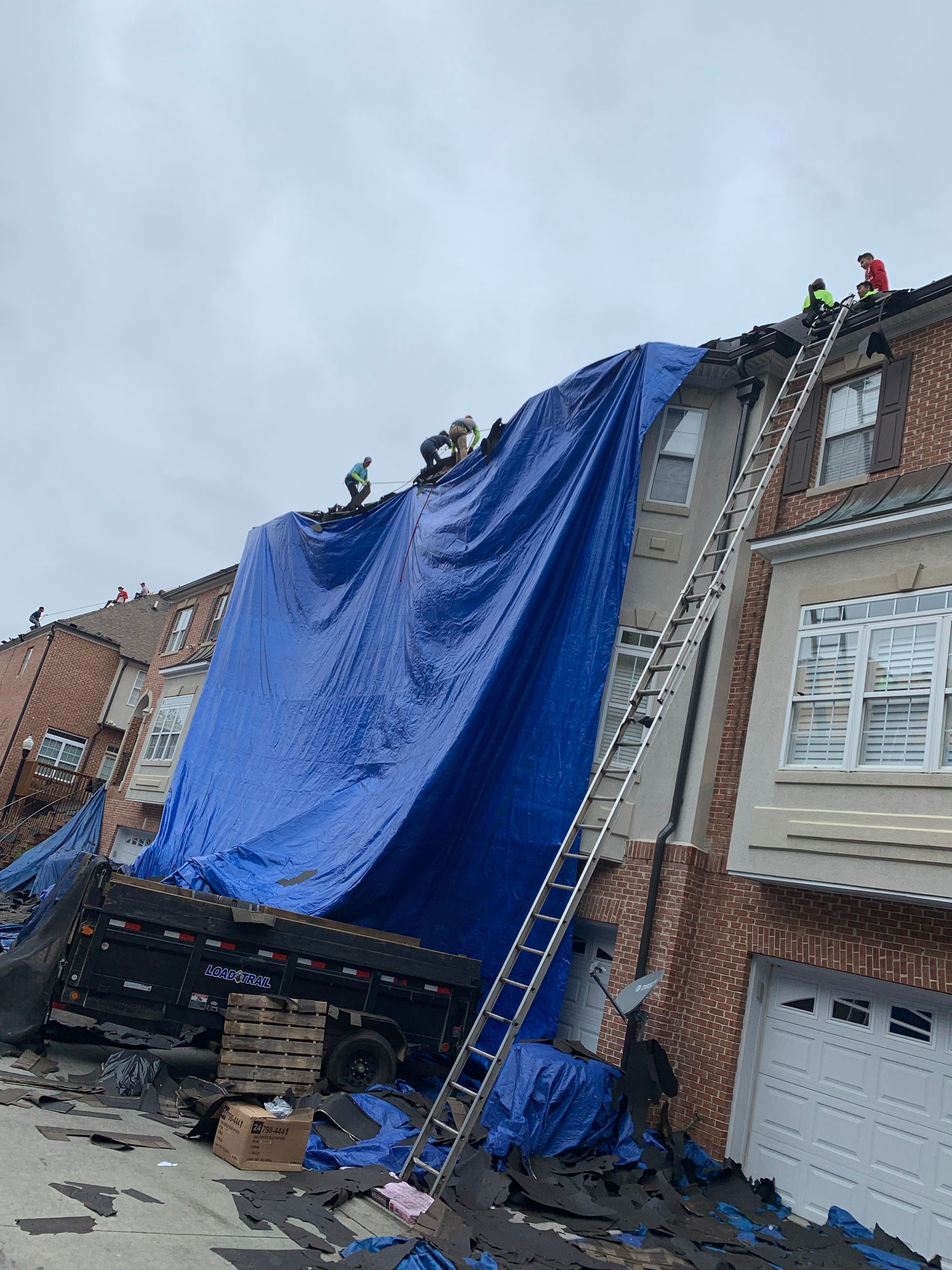 Roof covered in blue tarp with workers. Ladders and a truck are present. Overcast sky.