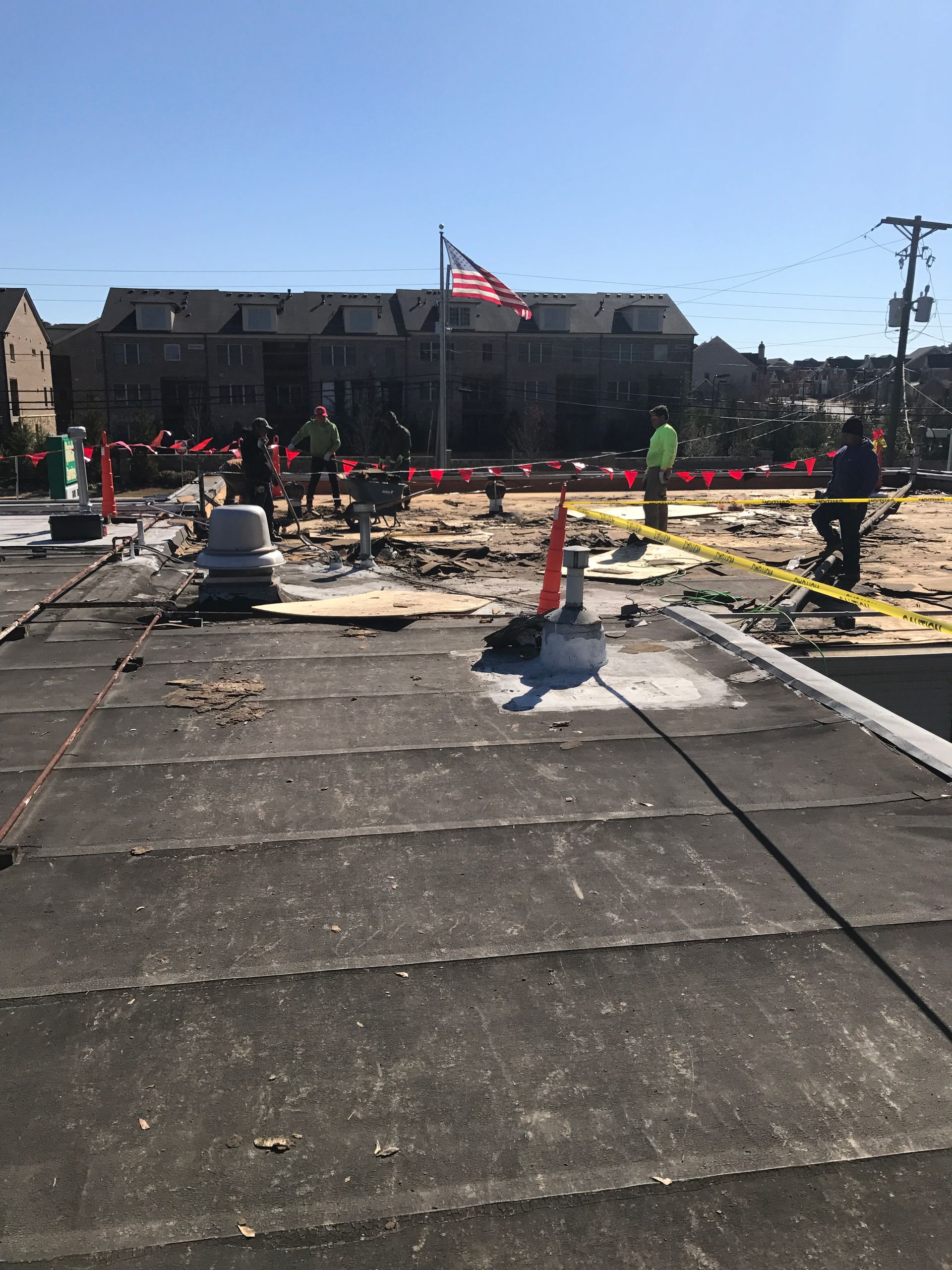 Construction workers on a flat roof; building in the background; caution tape; American flag.