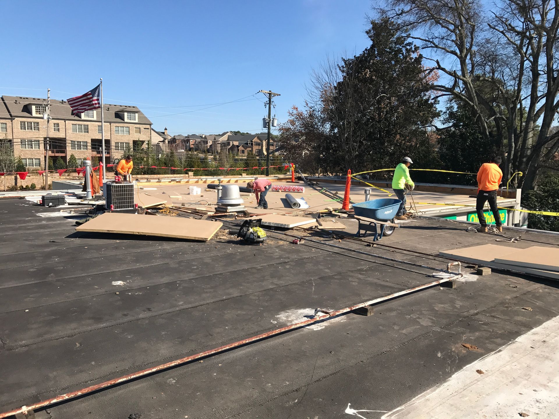 Construction workers on a flat rooftop. Safety barriers surround the work area. American flag in background.