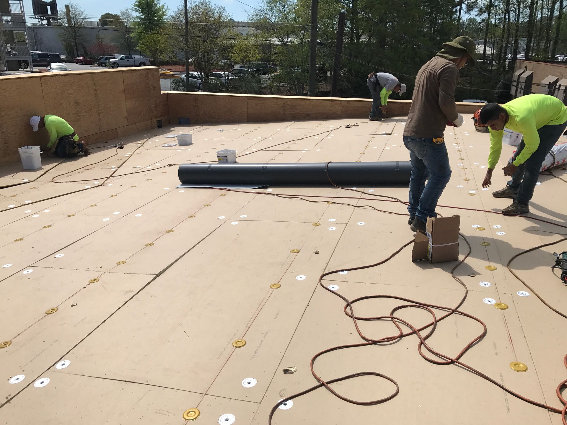 Construction workers installing roofing materials on a flat rooftop.  Men in safety vests work with tools and rolls of black material.