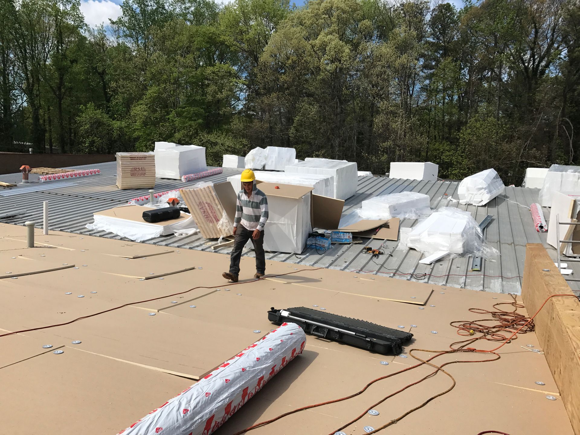 Construction worker on a roof surrounded by insulation materials and cardboard boxes. Trees in the background.