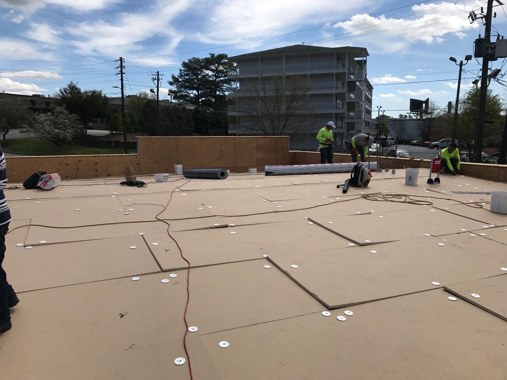 Construction workers on a rooftop, installing material. Beige surface, blue sky, and a building in the background.