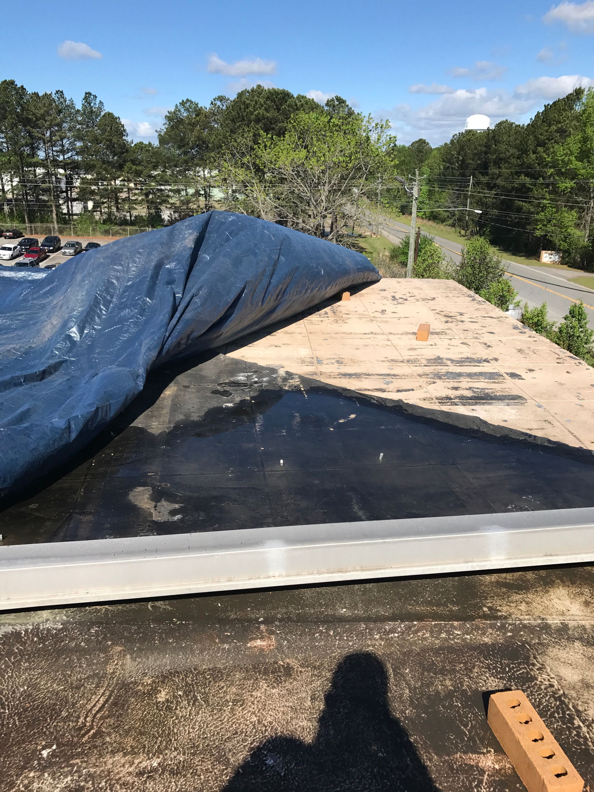 A blue tarp covers part of a damaged roof. Brick and trees are in the background, sunny sky.