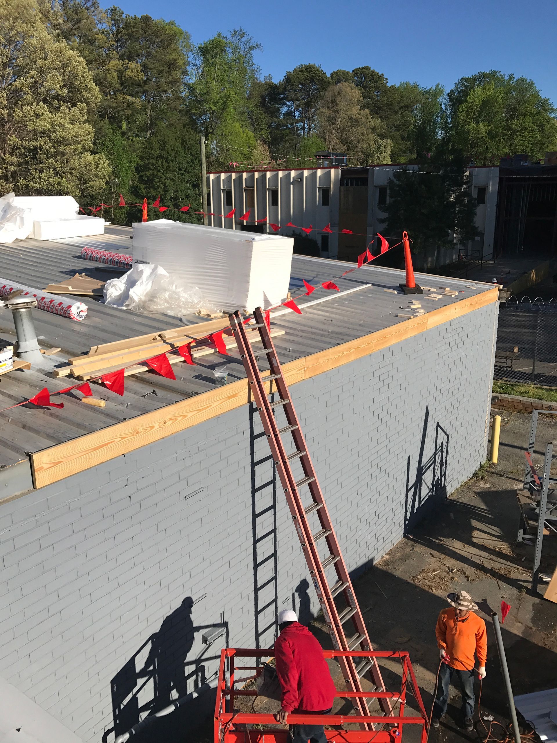 Construction workers on a gray building roof, using a lift and ladder, with safety flags.