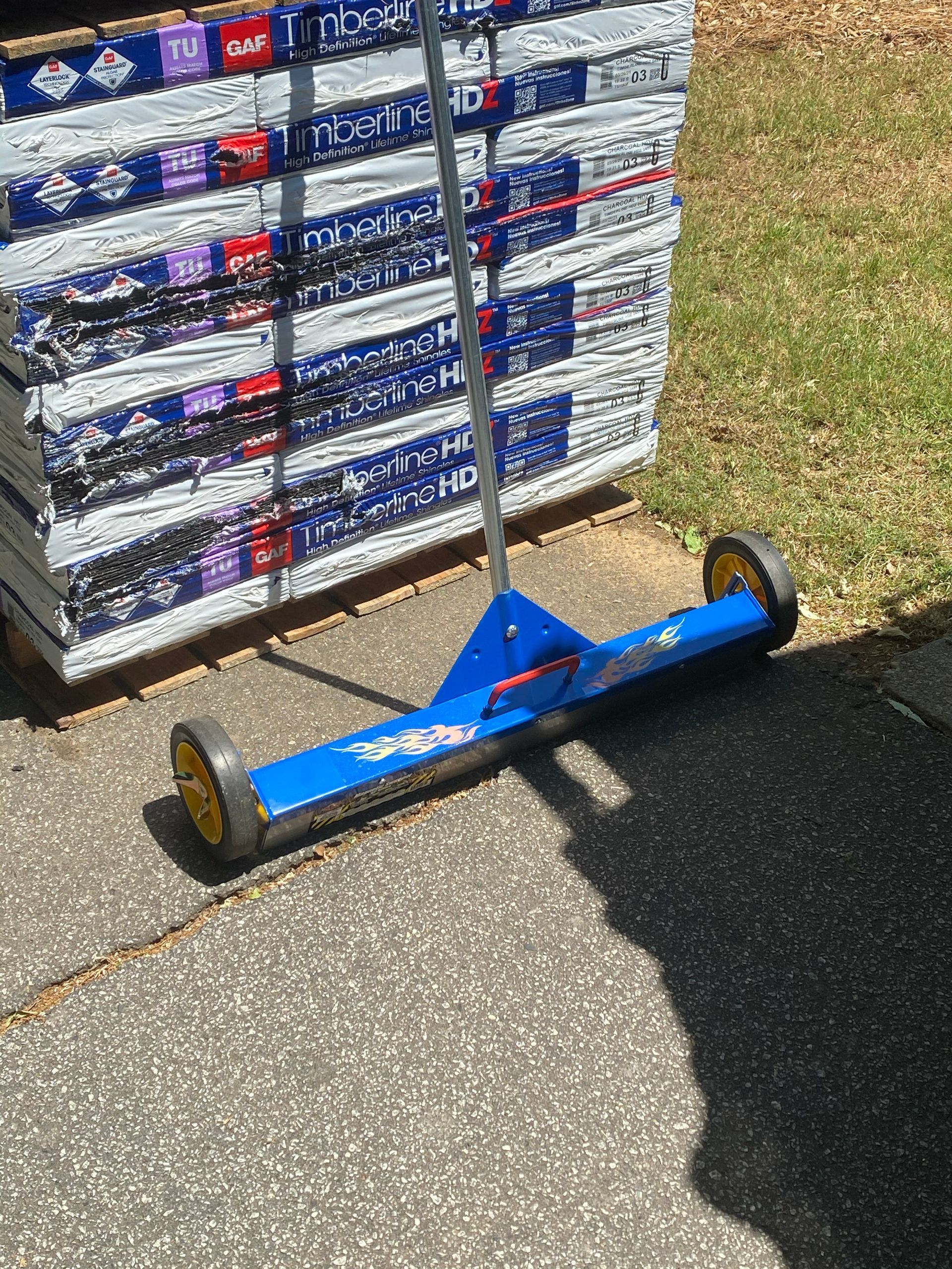 Blue magnetic sweeper on asphalt next to a stack of roofing shingles.