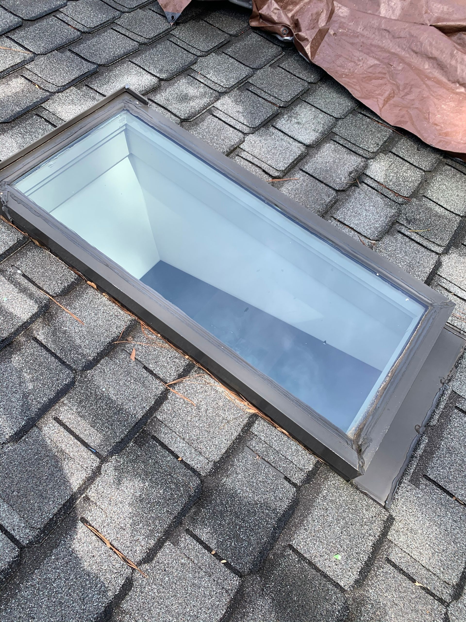 Skylight on a shingled roof, showing a rectangular opening with a metal frame.