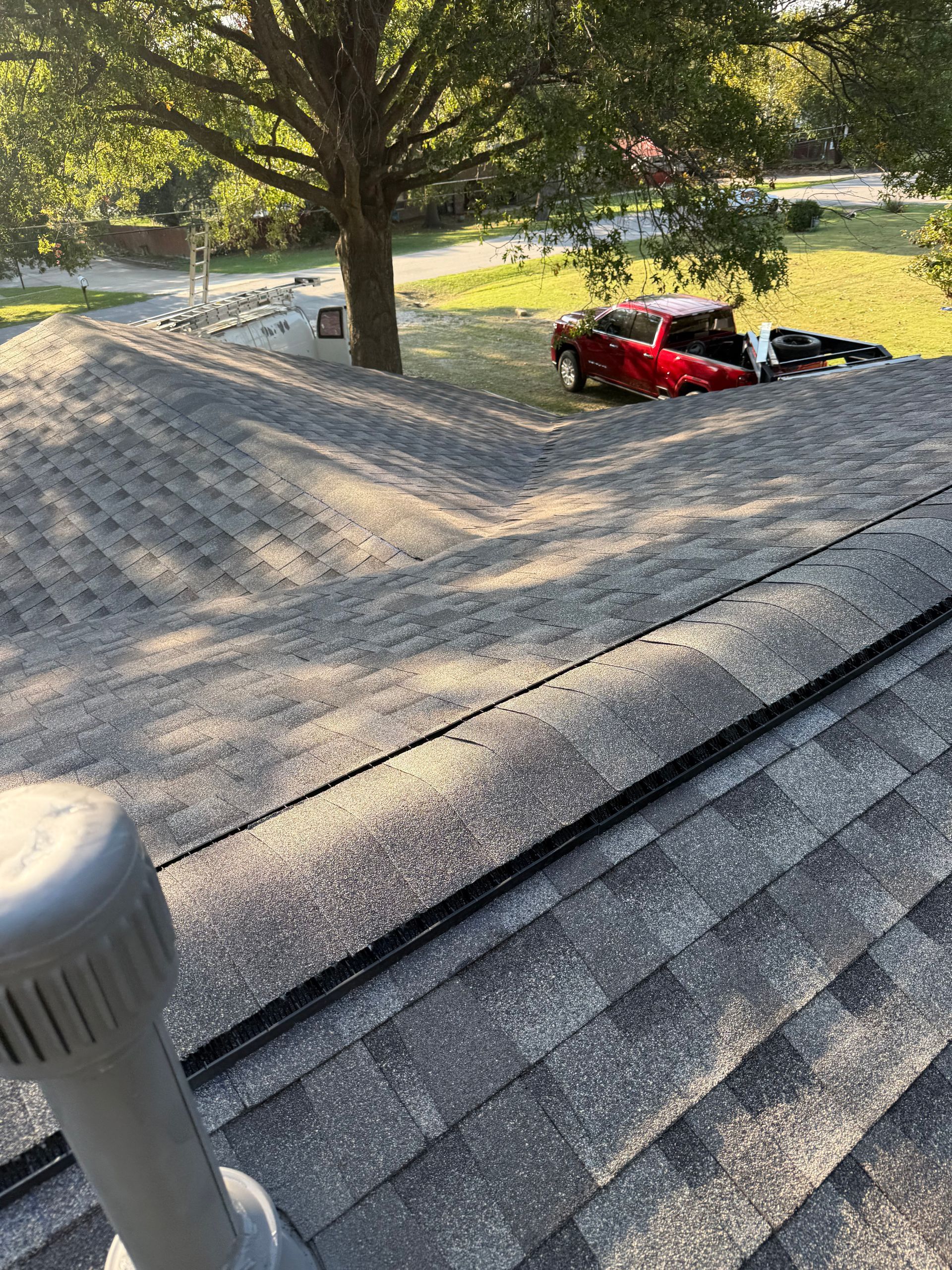 View of a shingled roof with a vent, trees, and a red truck in the distance on a sunny day.