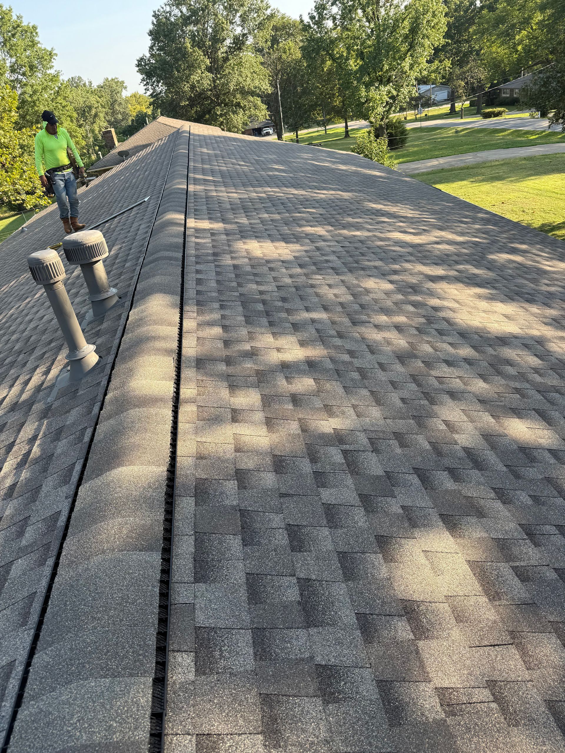 Person working on a weathered shingle roof, surrounded by trees and grass, with vents visible.
