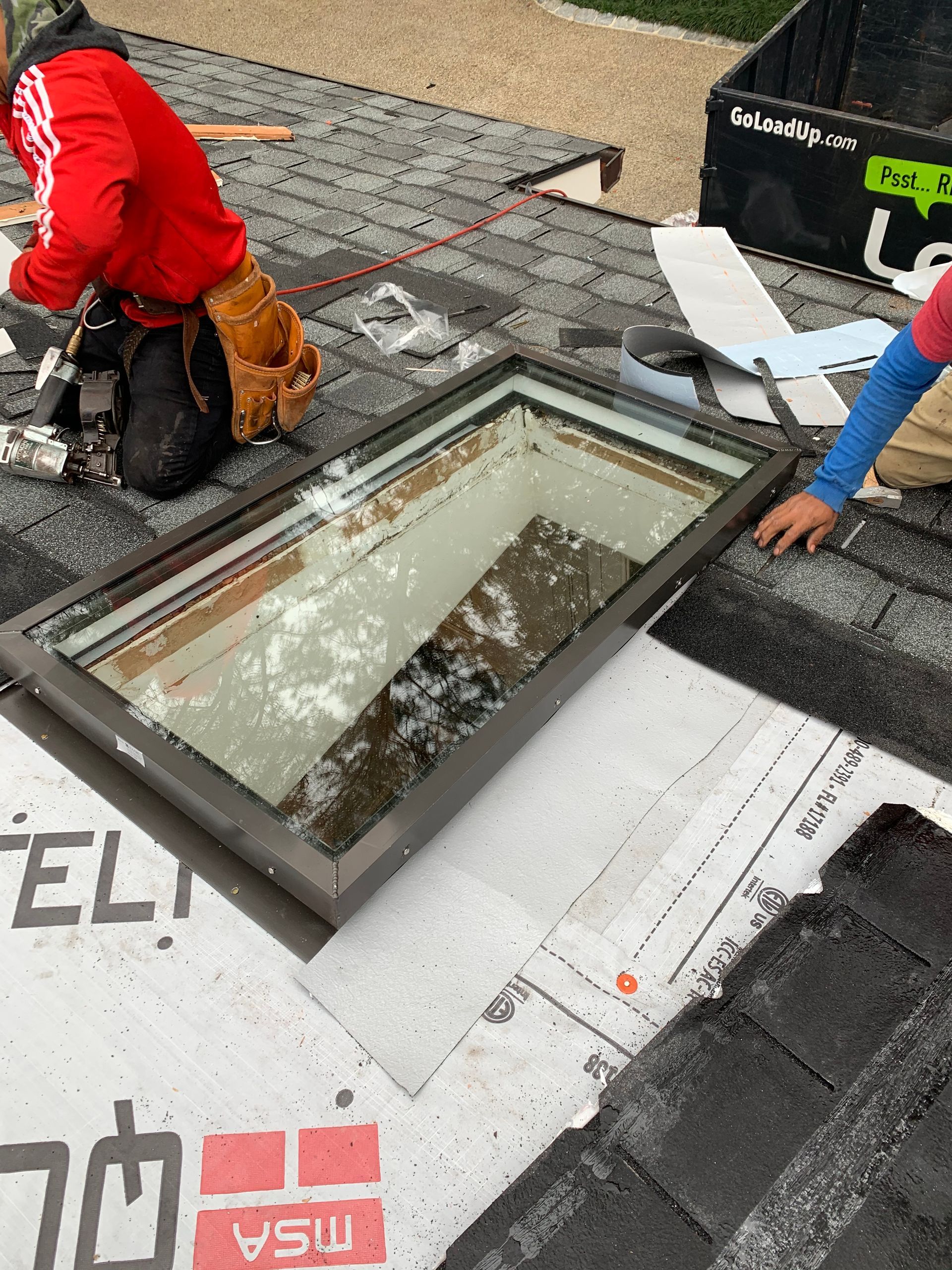Roofers installing a skylight on a rooftop. One worker in red kneels, another in blue reaches.