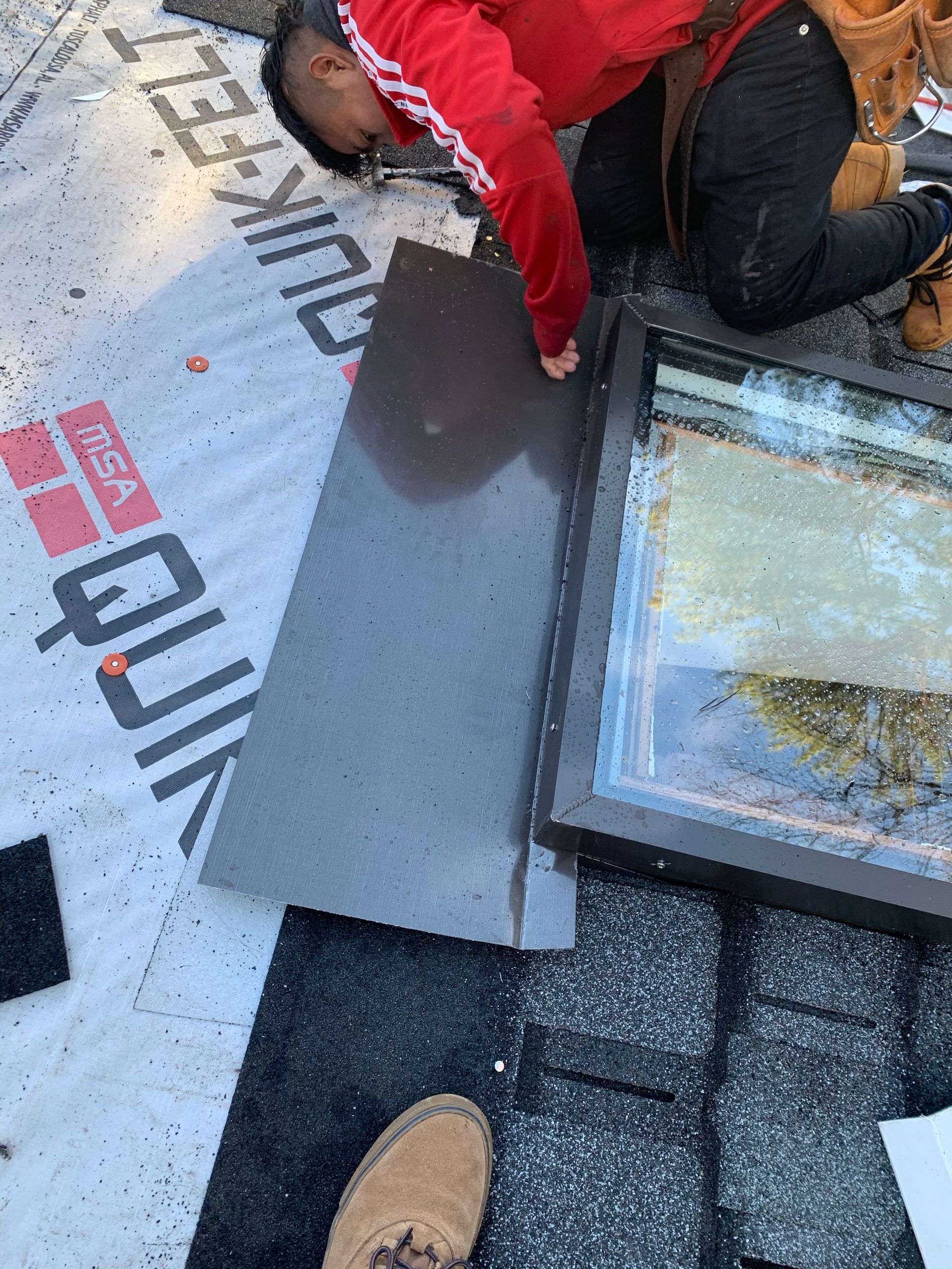 Roofer installing flashing around a skylight; brown metal flashing, red jacket, black shingles.