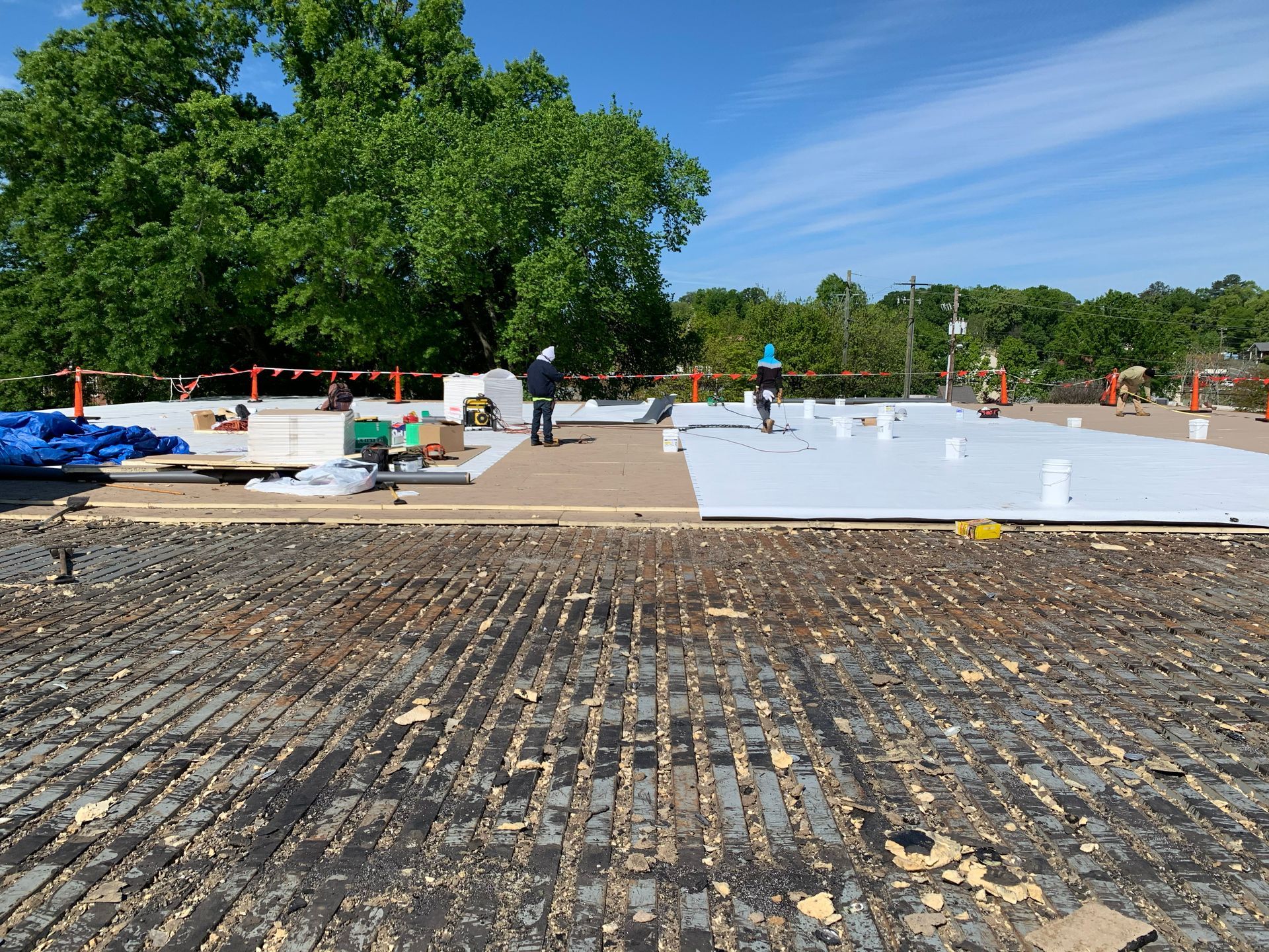 Workers on a rooftop install white panels. The roof is partially covered, with debris visible in foreground. Blue sky and trees.