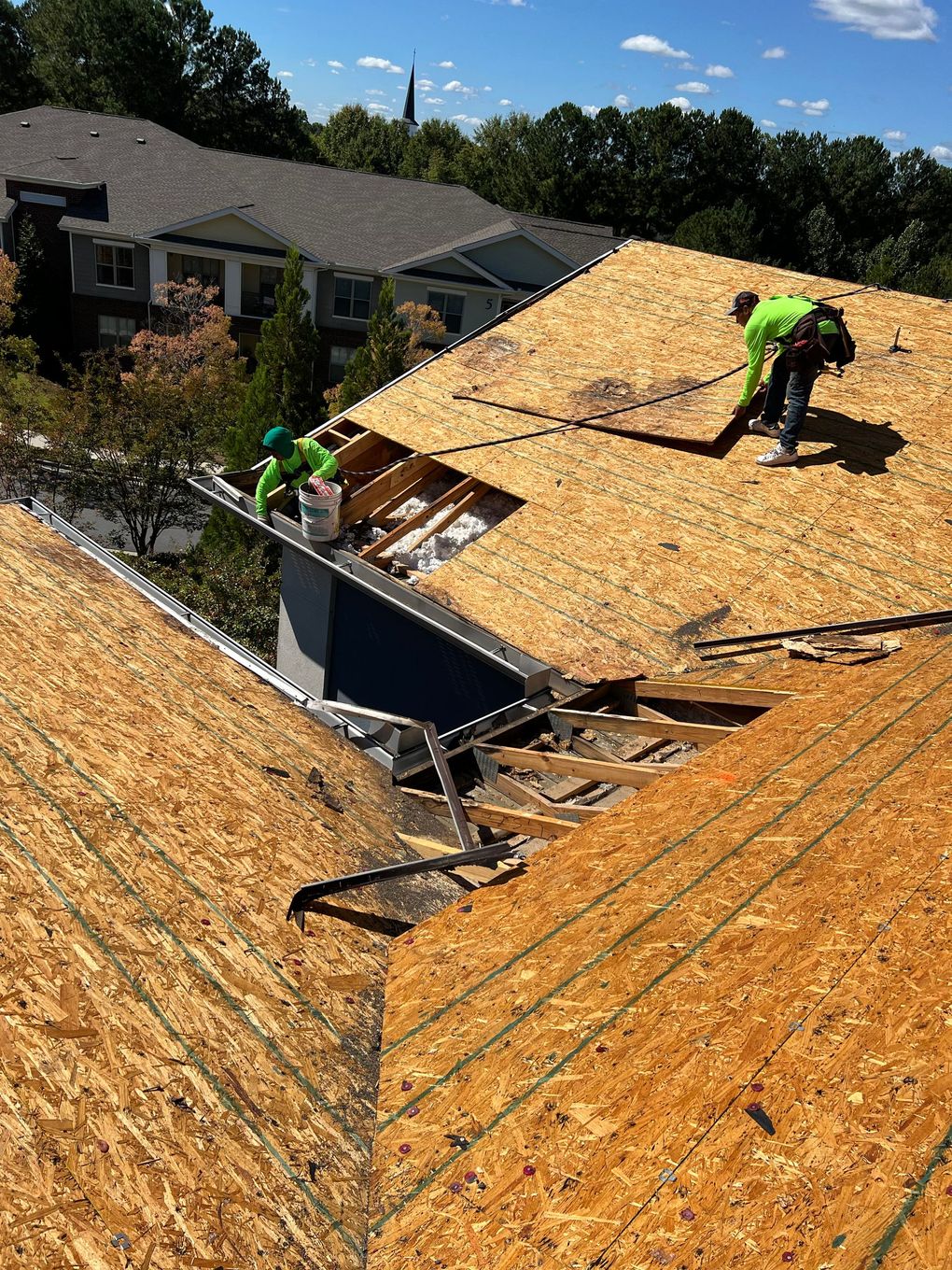 Two roofers in green gear on rooftop, working on new shingles; blue sky, homes in background.