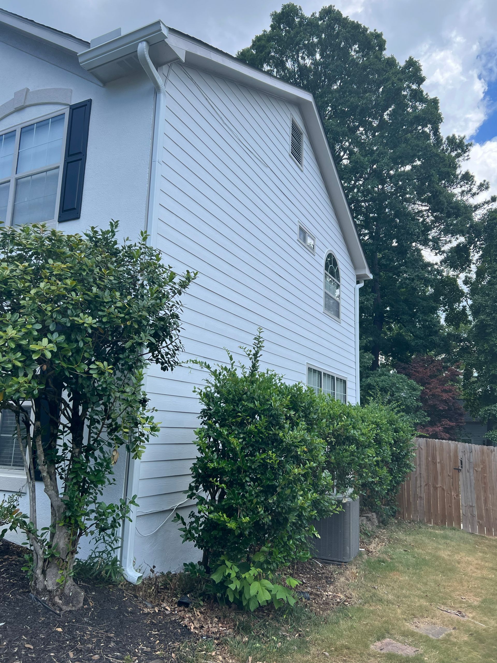 White two-story house with bushes in front and a wooden fence on the right side.