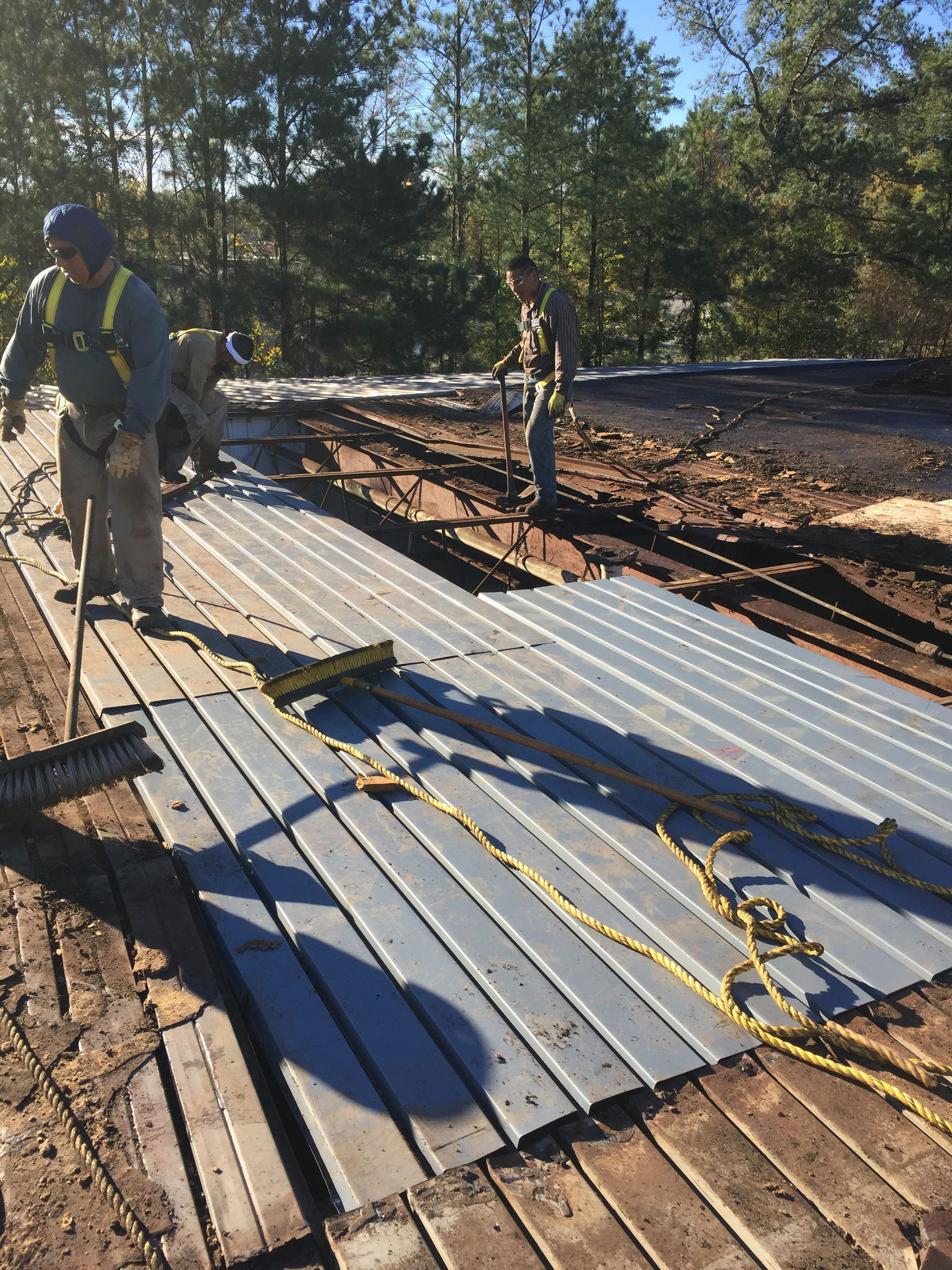 Construction workers installing metal sheeting on a roof. They are wearing safety gear and using tools in an outdoor setting.