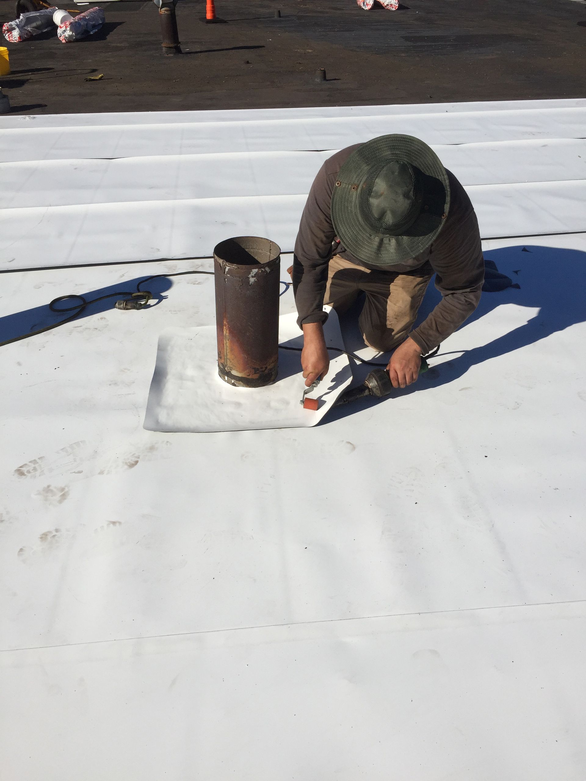 Person kneeling, applying sealant around a metal pipe on a white rooftop.
